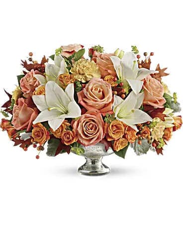 Autumn Shimmer Elegance - Celebrate the warmth and richness of the season with our Autumn Shimmer Elegance Centerpiece, a luxurious floral display designed to bring style, comfort, and sophistication to any fall setting. This breathtaking arrangement blends soft, romantic tones with vibrant autumn hues, creating a harmonious composition that instantly elevates your table décor or special occasion.  Lush peach and apricot roses take center stage, their velvety petals adding a touch of classic romance. They pair beautifully with crisp white lilies, whose radiant blooms bring freshness and elegance to the arrangement. Accents of gold-toned carnations, caramel spray roses, hypericum berries, and seasonal foliage enhance the design with texture, warmth, and autumn charm. Richly colored leaves and delicate greenery weave through the bouquet, capturing the cozy, golden glow of fall.  Presented in a stunning silver pedestal vase, this centerpiece exudes refined sophistication and timeless beauty. Its elevated design makes it the perfect focal point for dining tables, holiday gatherings, entryway décor, or heartfelt gifts for those who appreciate luxurious floral artistry.  The Autumn Shimmer Elegance Centerpiece is crafted to inspire gratitude, celebrate togetherness, and add a radiant seasonal touch to any space. It’s the ideal choice for Thanksgiving, anniversaries, or any moment that deserves something truly unforgettable.