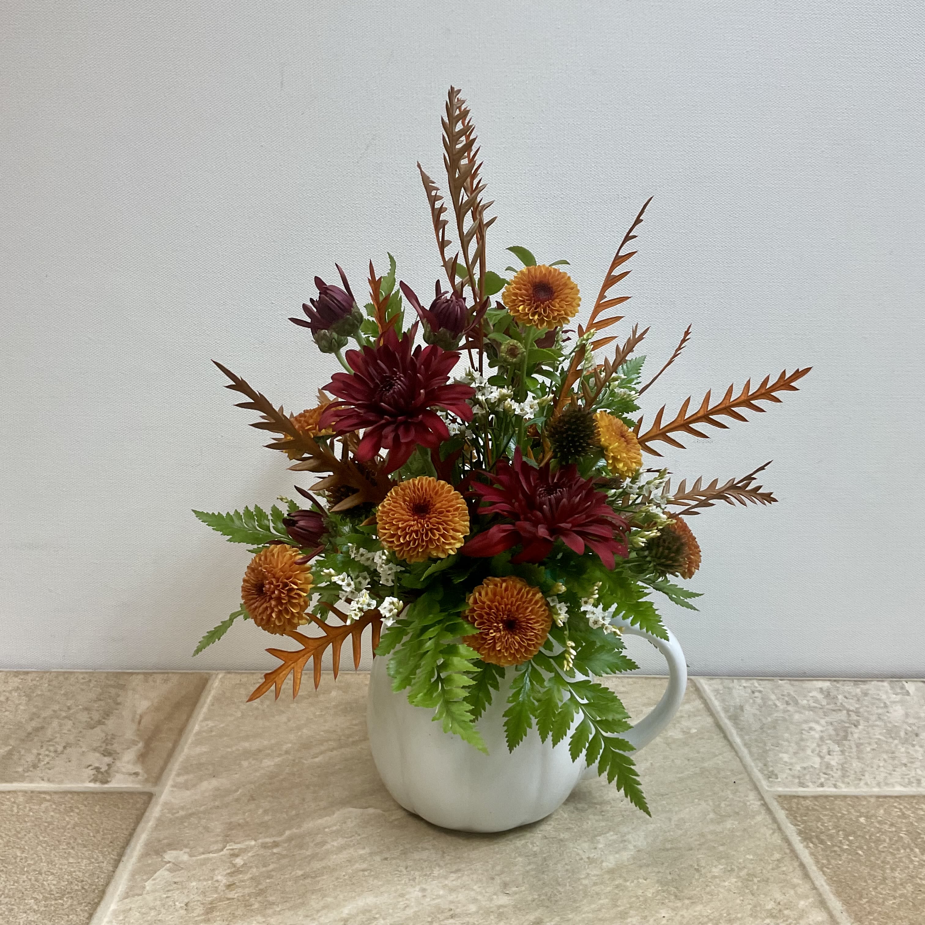 Cuppa Cozy Blooms - This arrangement has a warm, autumn-inspired look with rich, earthy tones. Deep burgundy chrysanthemums pair beautifully with burnt-orange pompom mums, while fresh green ferns and white filler flowers brighten and balance the bouquet. Wispy, copper-toned foliage adds texture and height, giving the piece a rustic yet elegant charm. The simple white ceramic mug base adds a cozy, homey touch, making it feel welcoming and seasonal. It measures 10” wide x 12” tall.