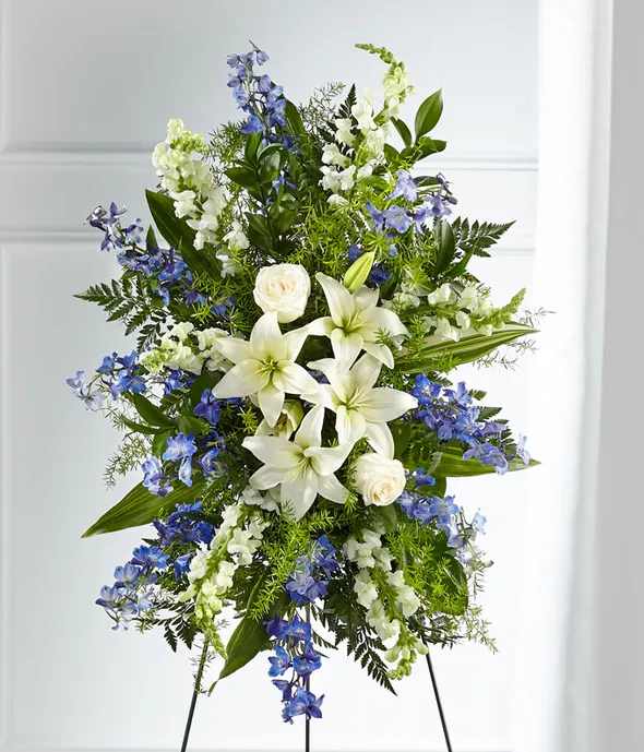 Faithful Friend Standing Spray - A collection of bold blue flowers shares a tribute that words cannot express. Designed with lilies, roses, delphinium and snapdragons our Faithful Friend Standing Spray is a grand gesture to express kindness and solace to a loved one while they are remembering the life they cherished so much.