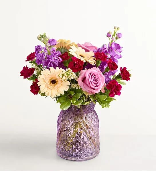 Alluring Beauty™ Bouquet - About Alluring Beauty™ Bouquet NEW From its variety of blooms to its eye-catching crescent shape, this is an out-of-the-ordinary bouquet for that out-of-the-ordinary someone. Roses, Gerbera daisies, and more, in buoyant peaches and purples, arrive lovingly arranged in a lavender vase with cottage charm