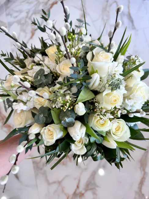 Elegant White Simplicity - This stunning handheld wedding bouquet features a timeless combination of white roses and lilies, accented with soft greenery. The white roses symbolize purity, innocence, and new beginnings, while the lilies represent rebirth and everlasting devotion. This is a classic and elegant bouquet that is perfect for any bride on her wedding day.