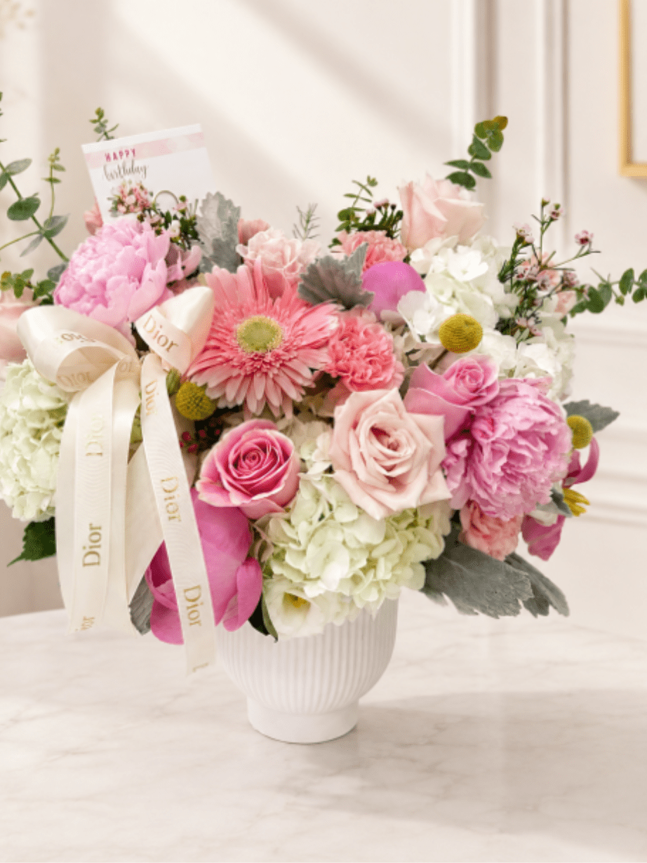 Blooming Grace - A delicate expression of beauty and elegance.  This sophisticated floral arrangement is beautifully designed in an elegant porcelain vase and features lush white hydrangeas, pink roses, pink Gerbera daisies, and 3 delicate pink peonies, complemented by eucalyptus and a curated selection of accent flowers that add texture, freshness, and graceful movement. 