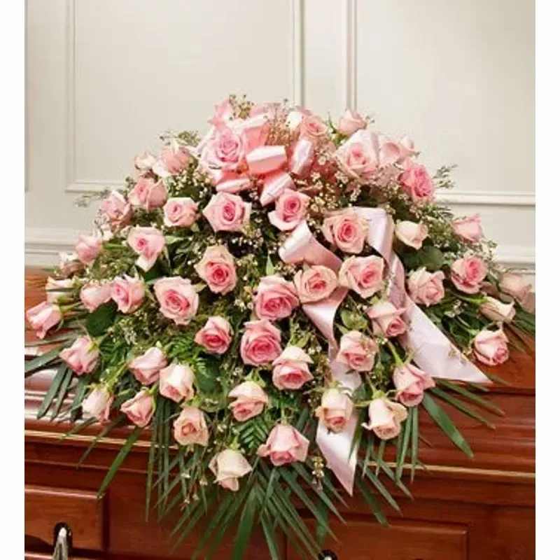 CASKET SPRAY - Pale pink roses arranged in a large, cascading casket spray create a gentle, solemn tribute for a funeral or memorial service. Designed as a casket arrangement, the soft pink blooms and rich dark-green foliage read as comforting and respectful-the kind of floral tribute that lets the person you're honoring feel remembered and cherished.  This spray features densely clustered pale pink roses at the center with trailing blooms that spill gracefully over the edge, accented throughout with white satin ribbons for a formal, refined touch. Long palm-like greenery extends beneath the flowers to give the piece its full, draping shape and traditional silhouette appropriate for placement atop a polished wooden casket.  Ideal for saying goodbye with dignity, this arrangement suits a close family member or dear friend and complements traditional funeral decor. Care is handled by the florist for delivery and placement; please confirm service details and timing when ordering to ensure the spray arrives for the ceremony.
