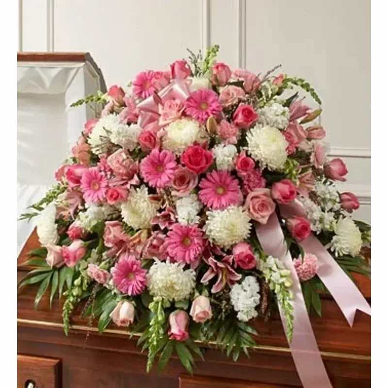 CASKET SPRAY222 - A layered casket spray in gentle pinks and whites that reads as both elegant and deeply sympathetic-ideal for a funeral or memorial service. This funeral flower arrangement combines roses, gerbera-style daisy blooms, carnations, chrysanthemums and lilies to create a soft, feminine tribute that conveys love and remembrance for the person you're honoring.  Built in full, tiered layers to sit across a casket, the spray features abundant rose heads and rounded white blossoms interspersed with bright pink daisies and carnations for contrast; a soft pink satin ribbon drapes across the front for a polished, respectful finish. The dense composition is arranged to present evenly from every angle and complements dark wood caskets with its luminous whites and warm pinks.  Choose this casket spray when you want the recipient to feel cherished and remembered-appropriate from close family members or dear friends as a signature piece at a service. Delivered ready to place on the casket, it's designed for immediate presentation and to hold shape throughout the ceremony; for ordering questions or delivery timing, please contact us so we can ensure timely, dignified delivery.