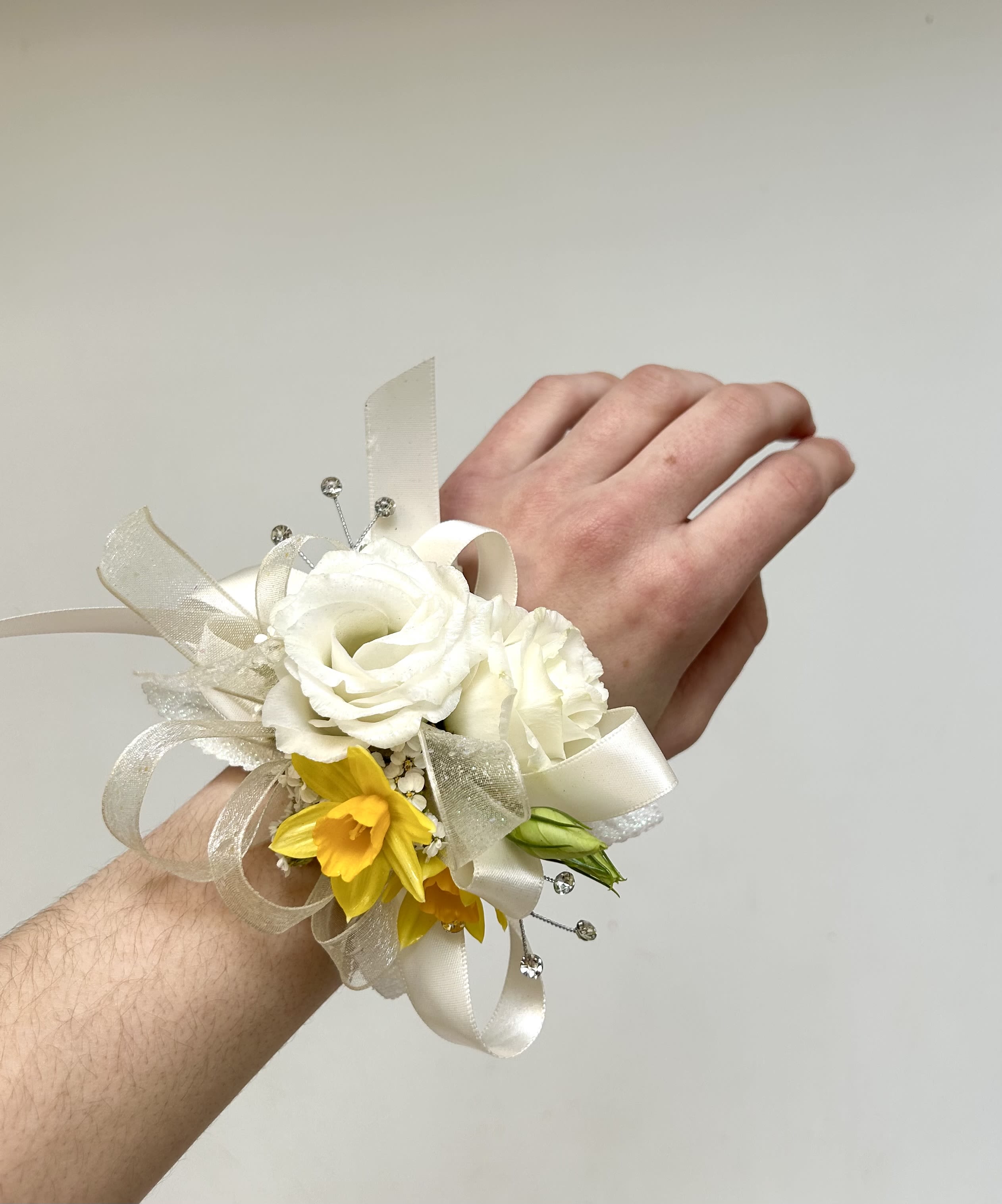 Yellow &amp; White Mixed Wrist Corsage - The compact design sits comfortably on a wrist band so the recipient can dance or mingle without fuss, while the soft ivory palette reads formal and timeless for photos and close-up moments.  This mixed-color corsage pairs full lisianthus blooms with bright daffodil accents, layered with ivory satin and sheer ribbon loops for classic texture. Small sparkling bead picks are tucked among the flowers to catch light and add delicate shimmer; finished on an adjustable wrist corsage band for a secure, comfortable fit. The arrangement's size is intentionally compact to complement rather than overpower formalwear.  Ideal for prom dates, bridesmaids, or anyone you want to honor on a special day, it sends a message of joyful elegance without being over the top. If you have specific flower requests, include them in the special instructions and we'll do our best to accommodate-please note substitutions may be necessary based on seasonal availability.