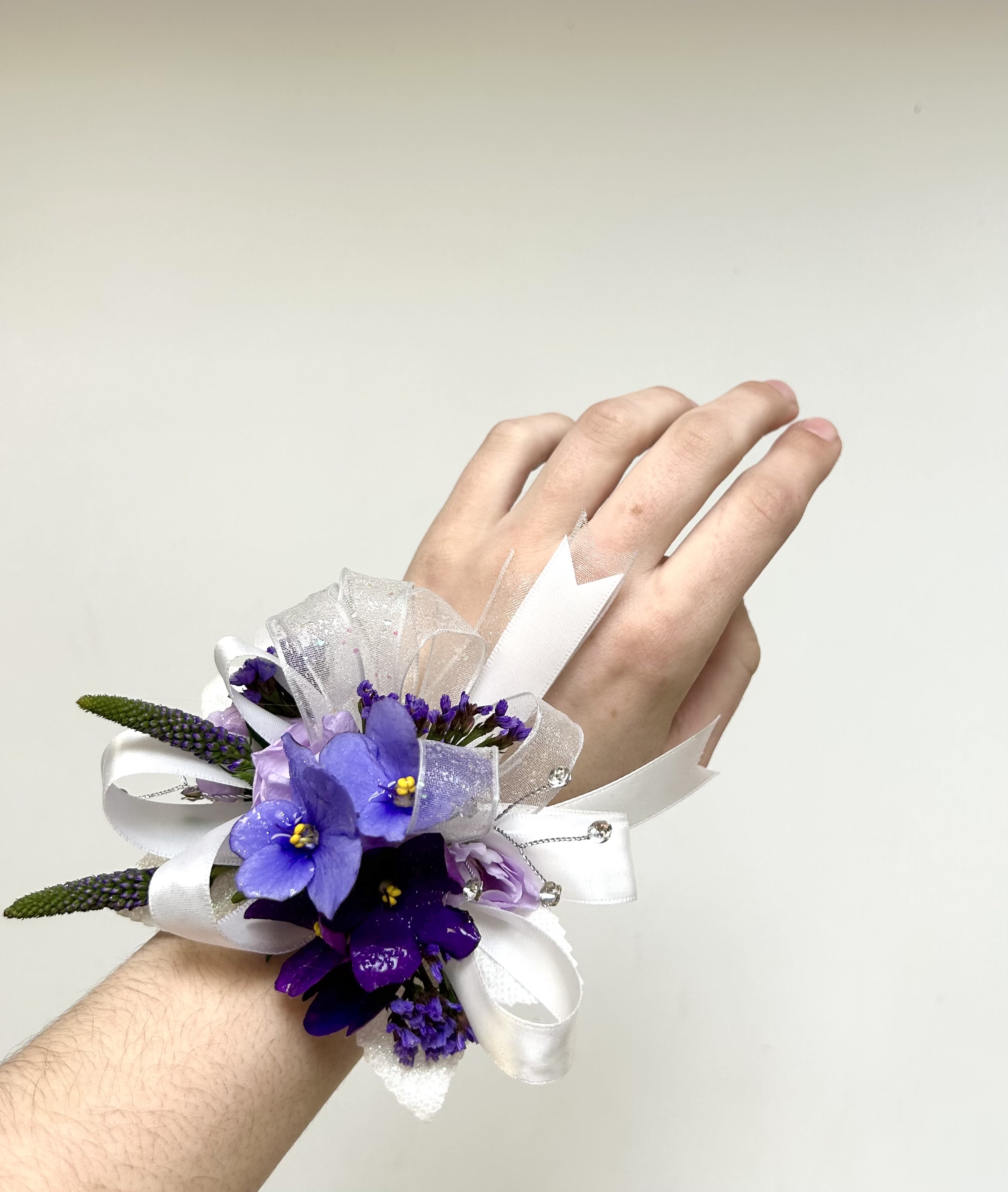 Purple Wrist Corsage - A cool-toned cluster of purple and lavender blooms creates an elegant, slightly dramatic wrist corsage perfect for prom or a formal wedding accessory. The compact arrangement sits neatly on the wrist.   Wrapped in layered white satin and sheer ribbon, finished with decorative beads, this corsage reads as refined and delicate against eveningwear. If you have a preference for specific flower varieties, please note them in special instructions and we will try our best to accommodate your request. Care: keep cool and mist lightly; best worn the day of the event.