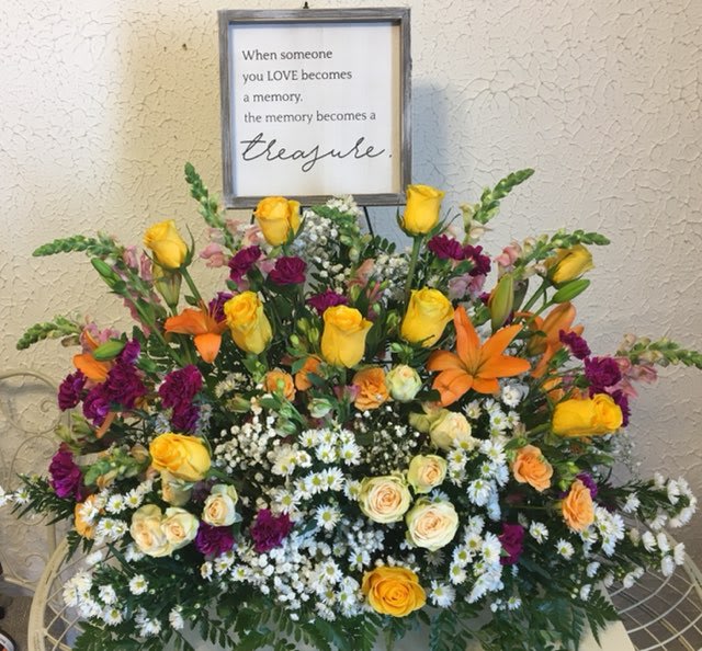 Large sympathy arrangement with yellow, orange, and purple flowers beneath a memorial sign