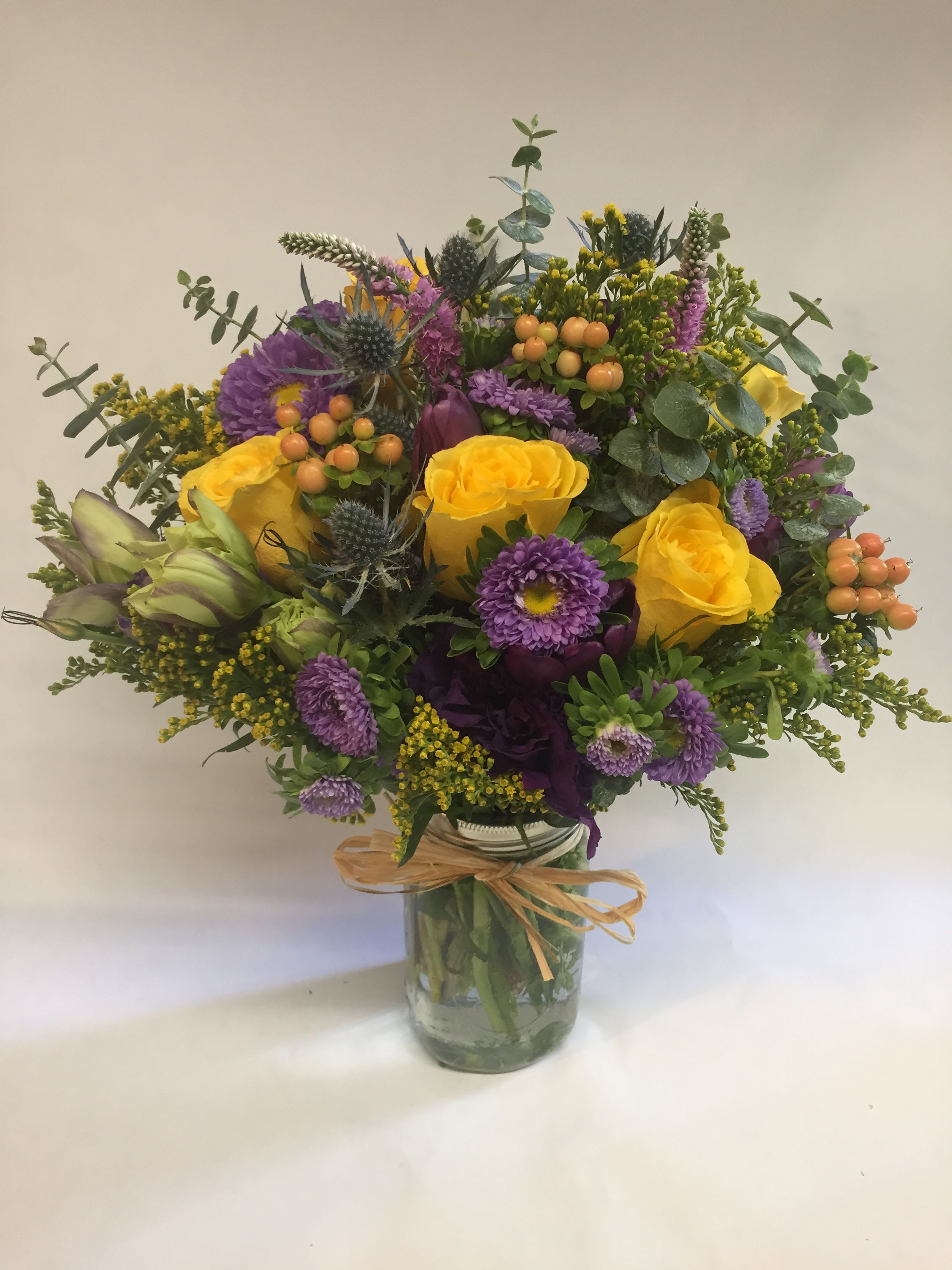 Bright yellow roses and purple asters arranged with berries in a mason jar tied with raffia