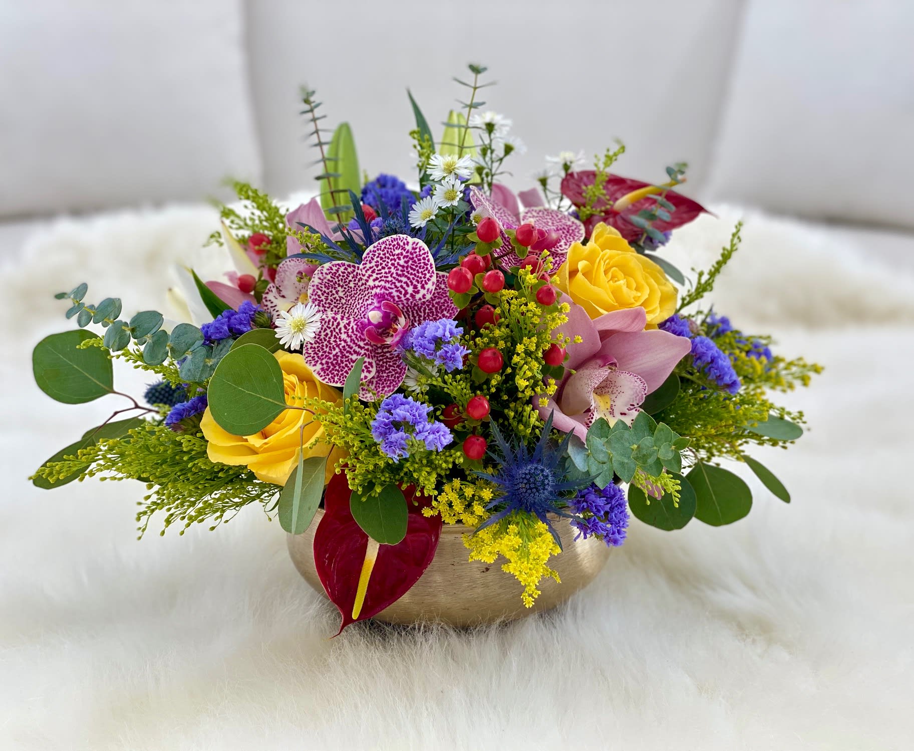 Joann ™ - This arrangement bursts with vibrant color and texture, blending sunny yellow roses, speckled orchids, and soft pink cymbidium orchids with striking blue thistle and statice. Red hypericum berries and anthuriums add bold contrast, while eucalyptus and golden fillers complete the design. Presented in a chic gold vase, it’s a lively, artistic centerpiece full of energy and elegance.