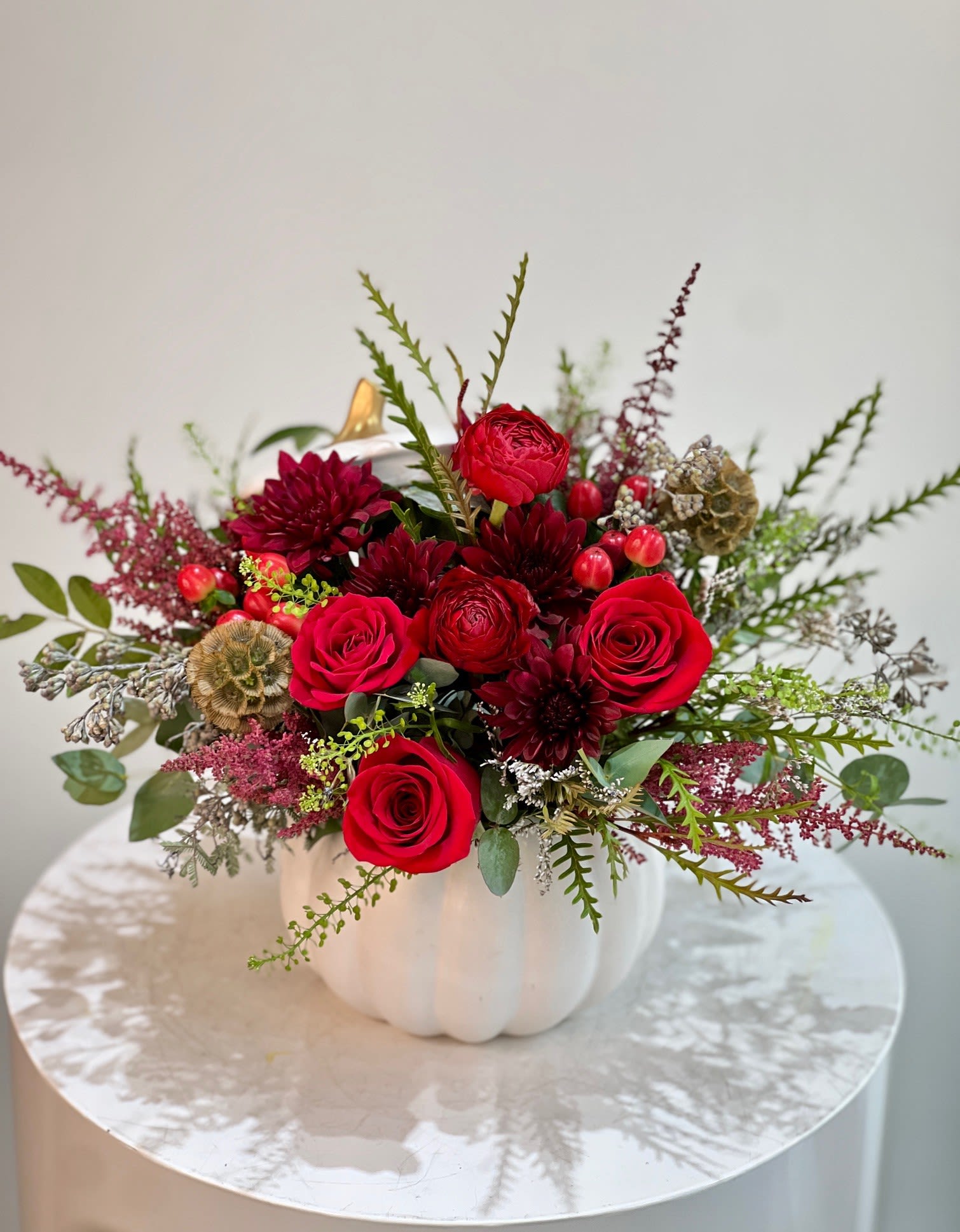 Ceramic Pumpkin with fall flowers/ red flowers ™ - This bold autumn-inspired arrangement bursts with romance and warmth. Deep red roses, ranunculus, and burgundy dahlias are beautifully complemented by textured greenery, berry accents, and dried pods—all elegantly arranged in a white pumpkin vase topped with a golden stem. Perfect for seasonal celebrations or as a statement centerpiece, this design captures the essence of fall with a luxurious, modern twist.