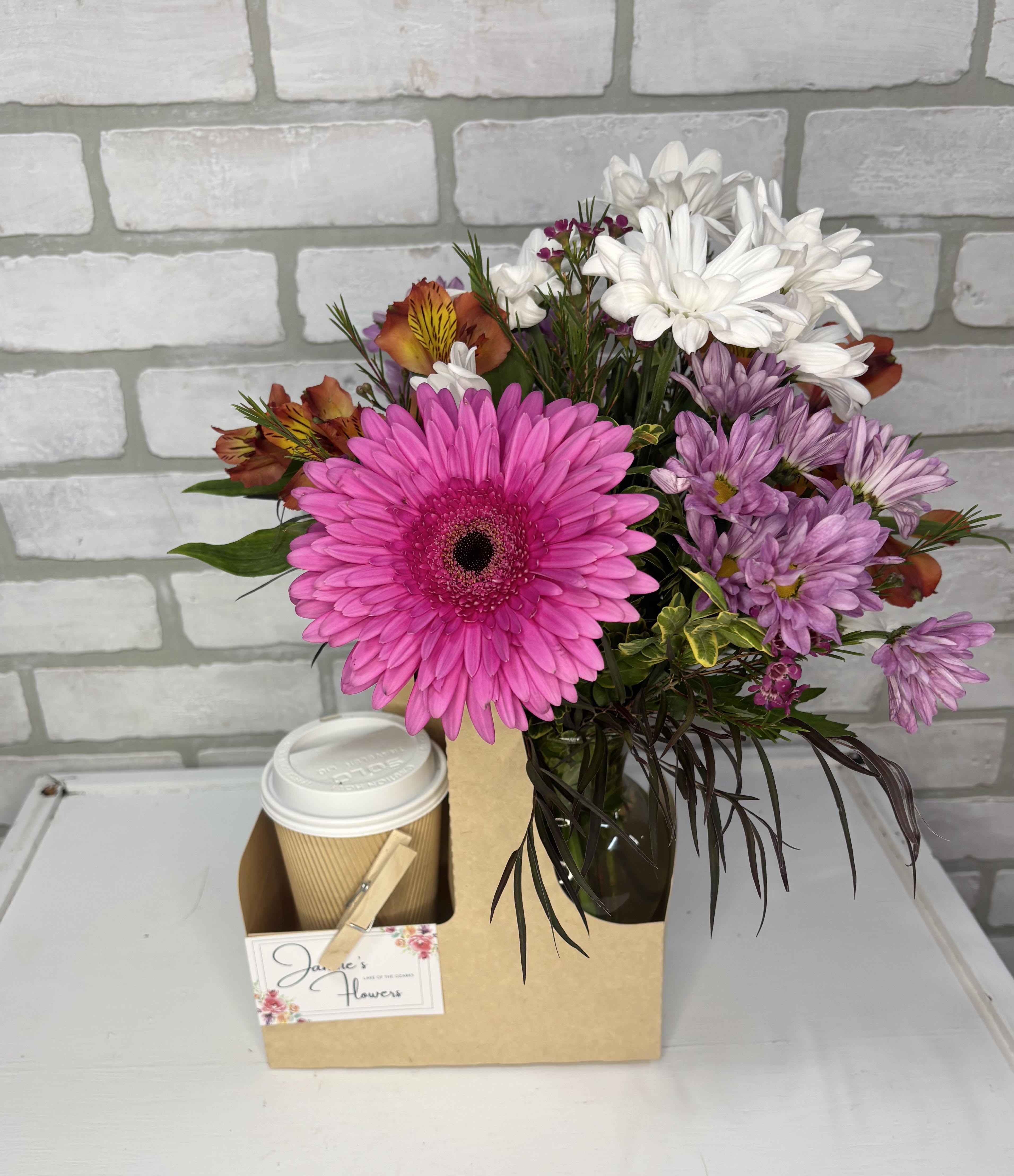 Mixed bouquet with bright pink gerbera daisy in glass vase next to a to-go coffee cup in a cardboard caddy.