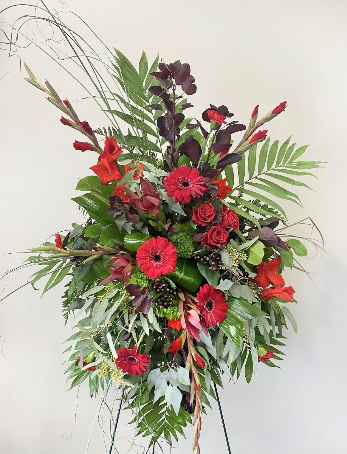 Tall standing spray of red gerbera daisies and roses with dramatic greenery on an easel.