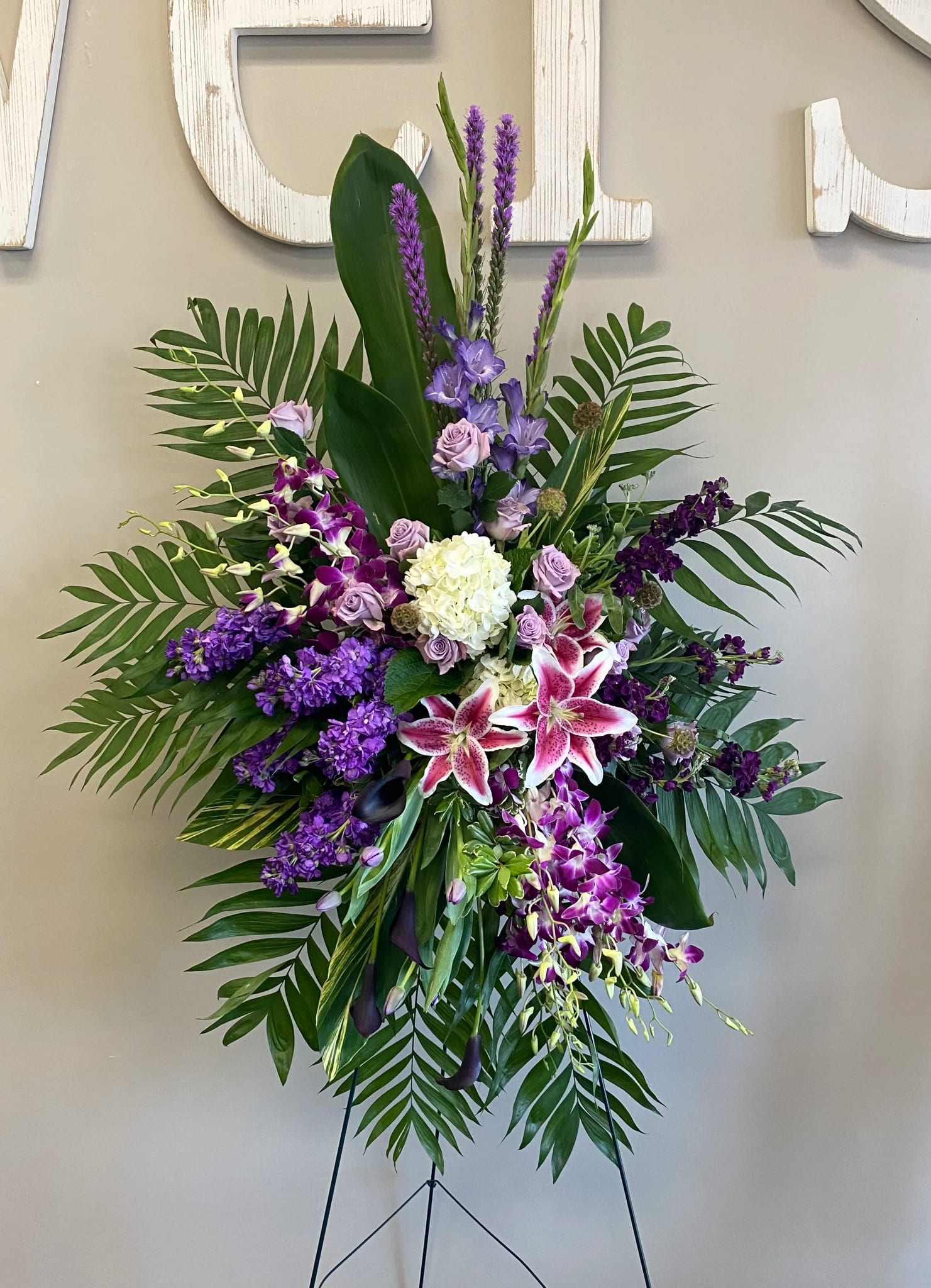 Tropical Purple ES-11 (Pictured as Deluxe) - *DELUXE OPTION PICTURED*  Elegant and expressive, this standing spray showcases a rich palette of purples, lavenders, and soft white. Featuring fragrant stargazer lilies, orchids, stock, roses, liatris, and hydrangea, the arrangement is styled with lush greenery and tropical leaves for a striking presentation. Displayed on an easel (stand included), this piece offers a graceful and heartfelt tribute, perfect for honoring someone with a vibrant spirit and a deep sense of beauty.