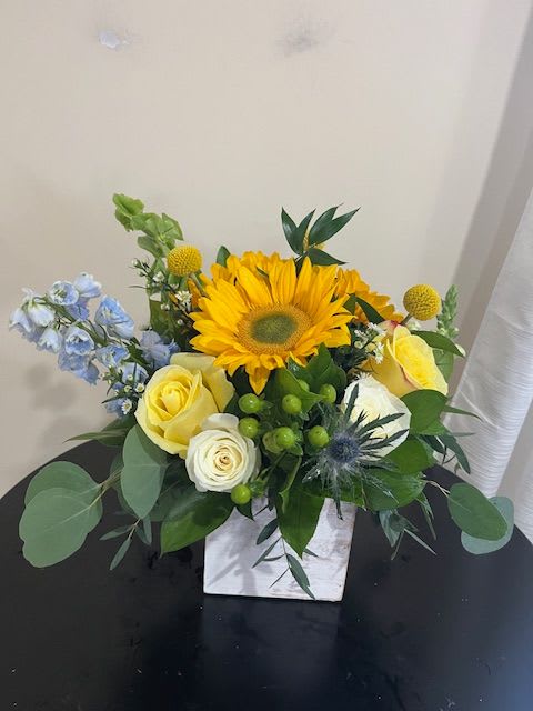 Bright sunflower and yellow roses with blue accents in a white cube vase