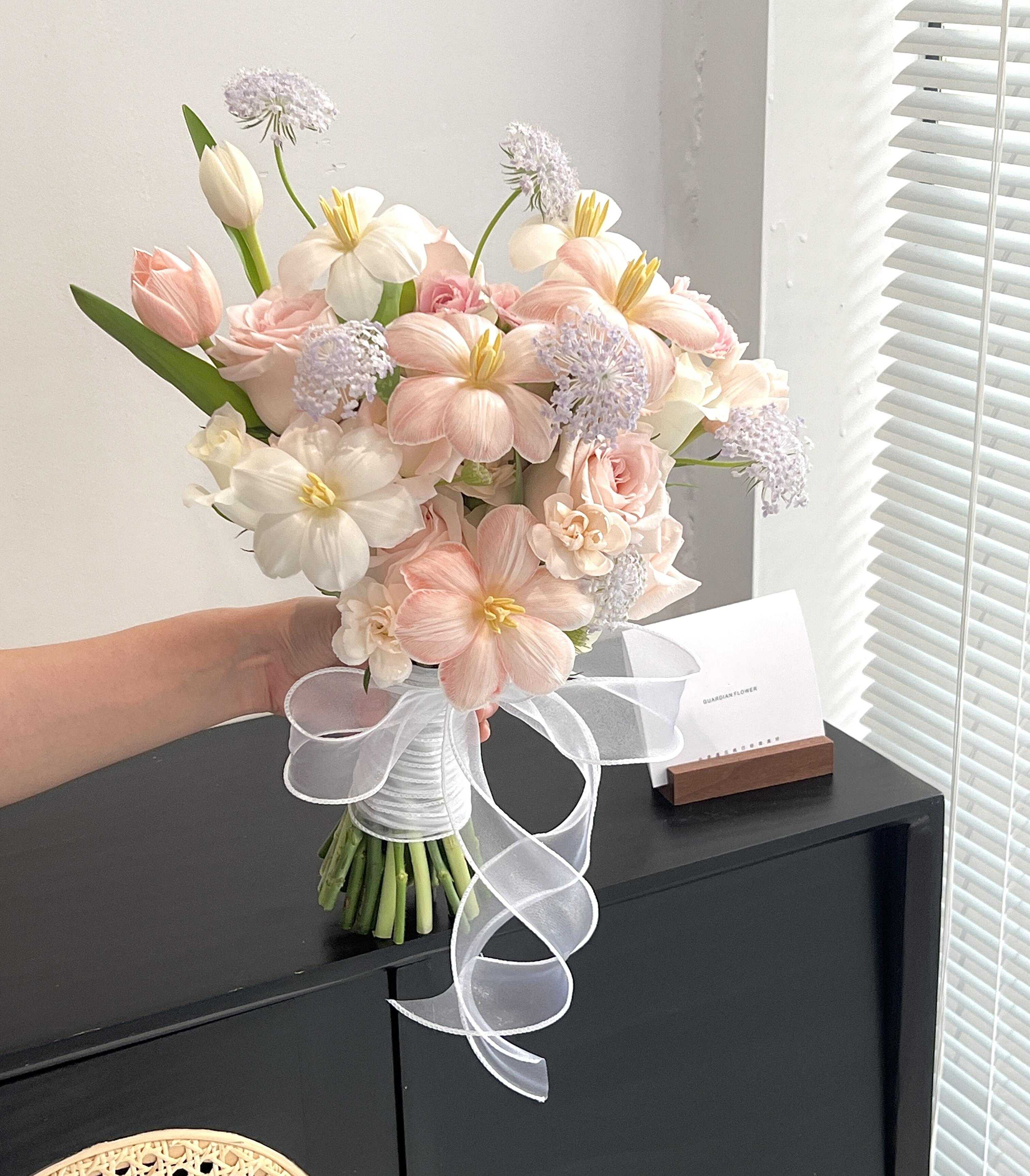 Handheld bouquet of pale pink and white flowers tied with a white ribbon