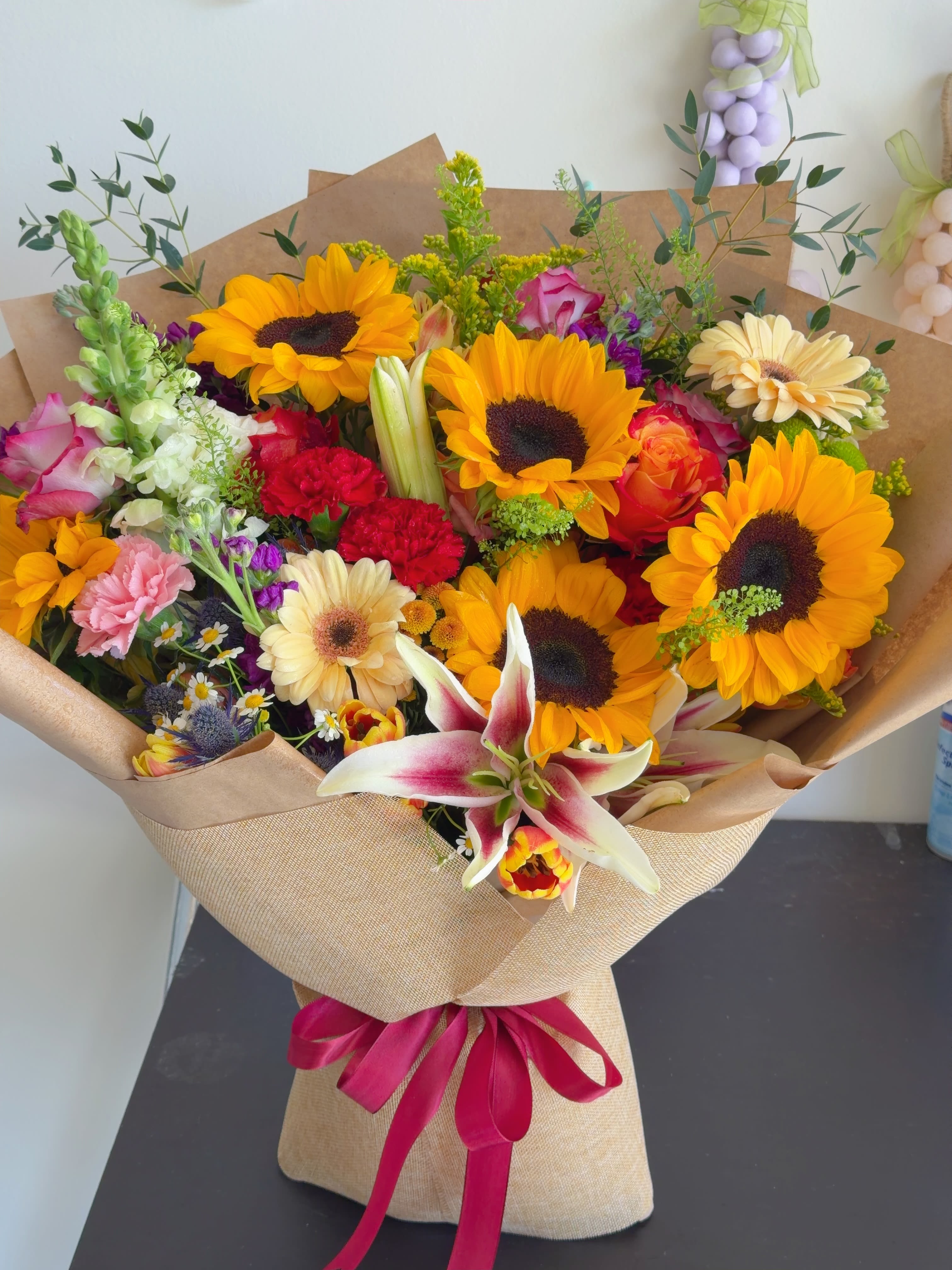 Radiant Summer Bloom  - Radiant Summer Bloom is a vibrant bouquet featuring cheerful sunflowers, elegant lilies, roses, and seasonal flowers, beautifully wrapped in signature Korean-style paper. Bright, joyful, and full of color, it’s perfect for birthdays, celebrations, congratulations, thank-you gifts, or simply making someone’s day brighter.  Available in Standard, Deluxe, and Premium sizes. Flower varieties and wrapping paper may vary slightly based on seasonal availability while maintaining the same beautiful style and color palette. Each bouquet is handcrafted with fresh flowers.