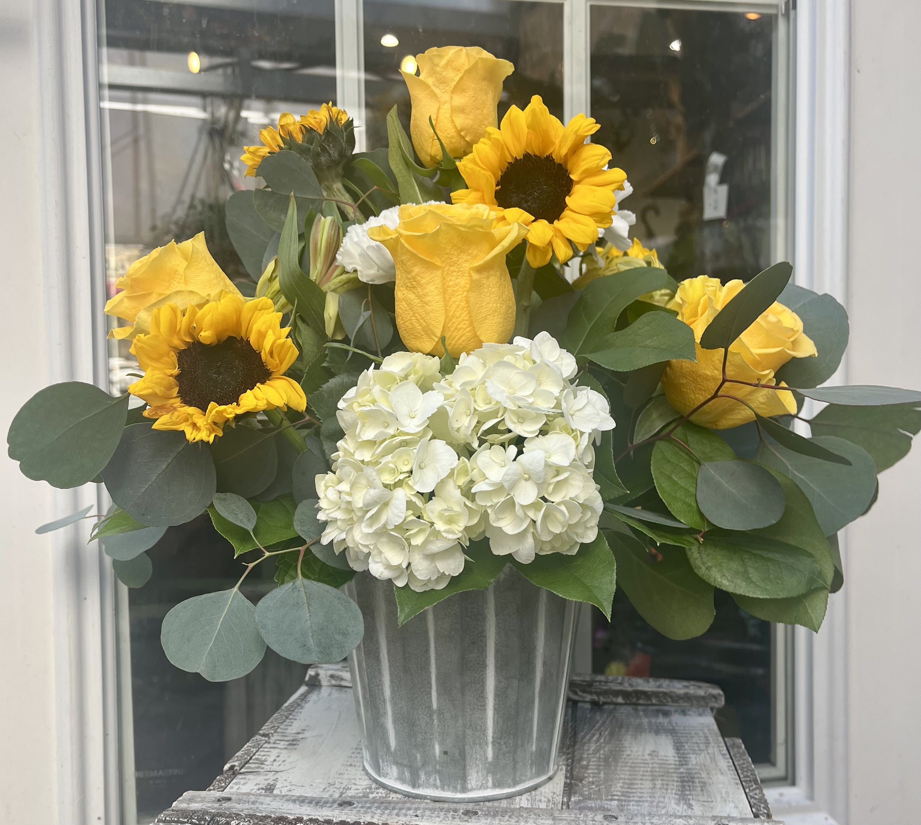 Yellow roses, sunflowers, and white hydrangeas arranged in a metal bucket-style vase