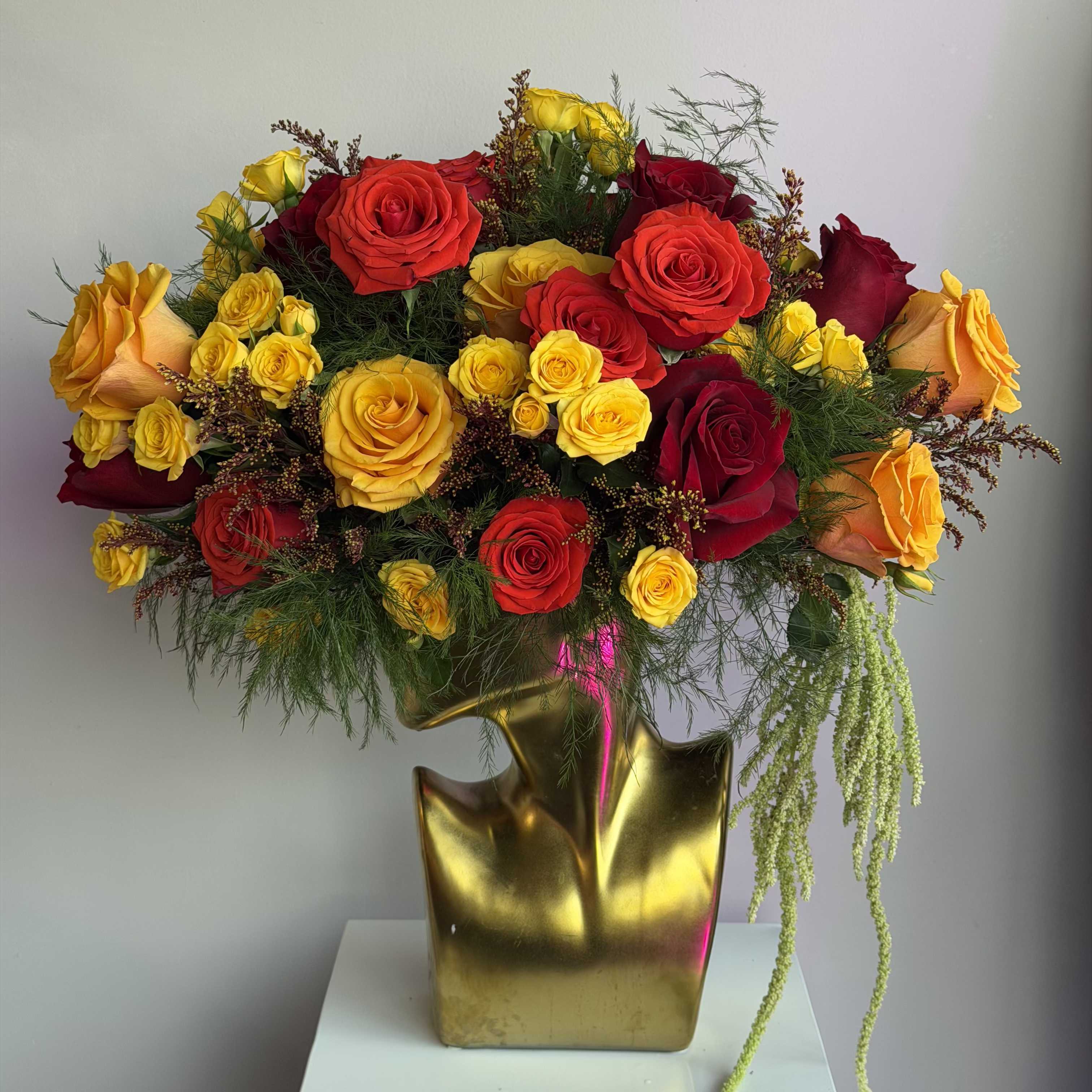 Tall arrangement of red, yellow, and orange roses in a sculptural gold vase