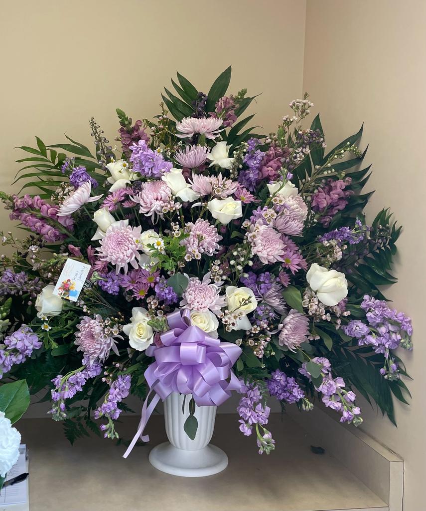 Large lavender and white floral arrangement in a white urn with a purple bow
