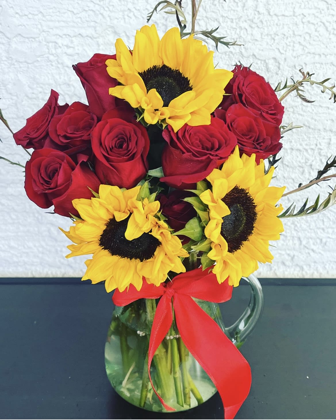Red roses and yellow sunflowers in a clear glass pitcher with a red ribbon
