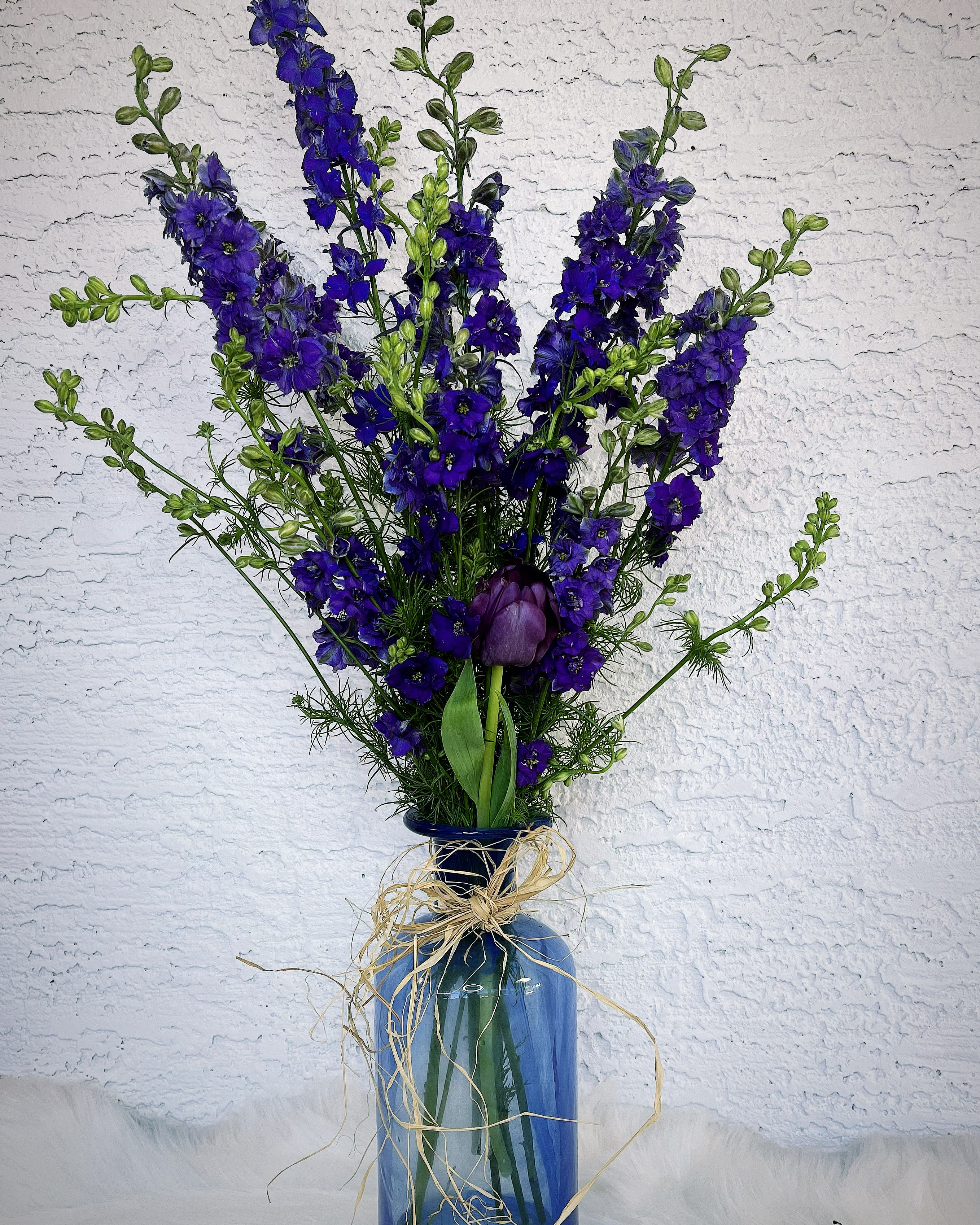 Tall blue floral arrangement in a glass vase with raffia bow