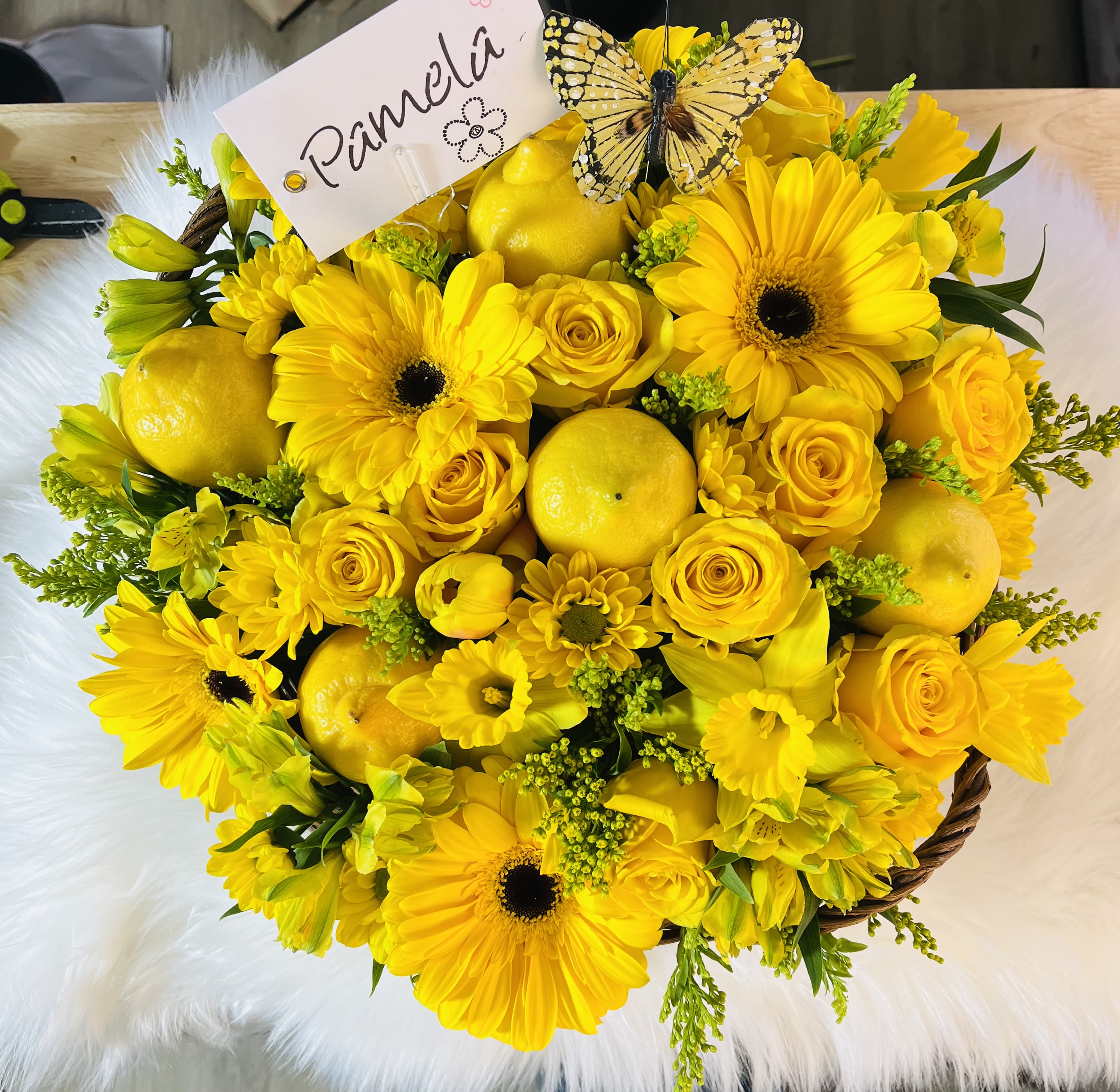 Round yellow flower basket with gerbera daisies, roses, lemons, and a name card topped with a butterfly.