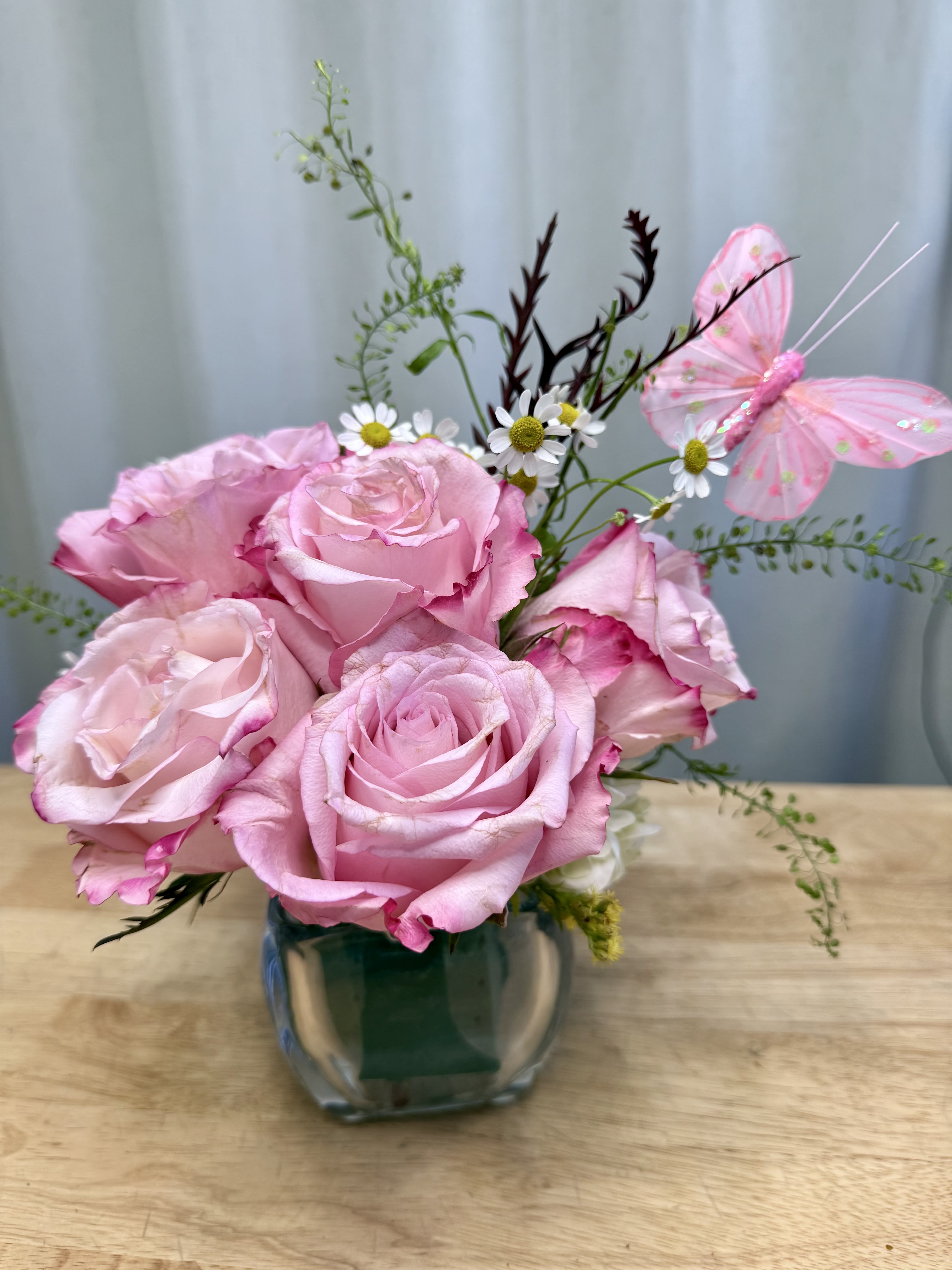 Pink roses in a glass vase with small white daisies and a pink butterfly decoration.