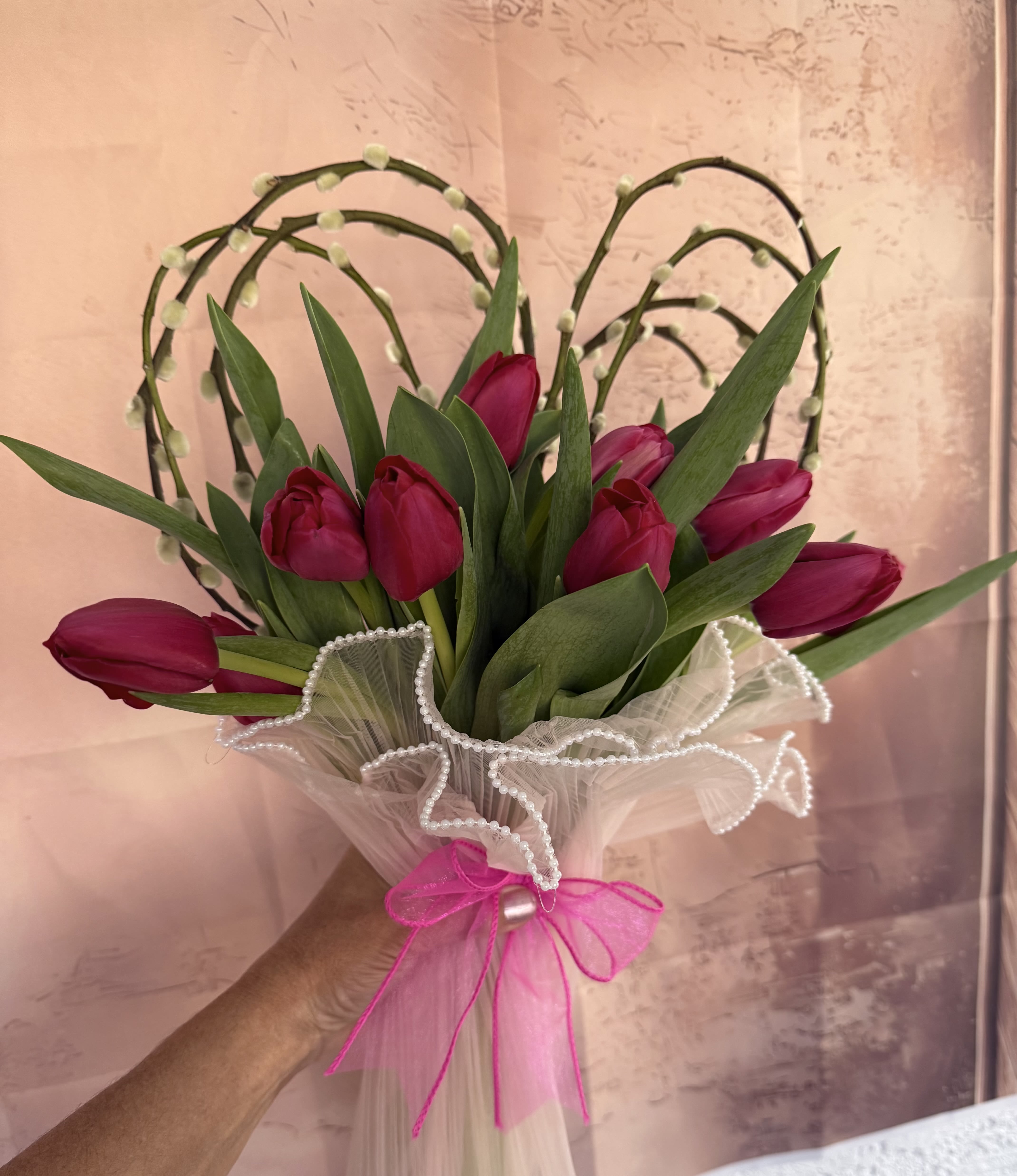 Blushing Heart Tulip Bouquet - A sweet and elegant bouquet designed to make hearts melt. This arrangement features vibrant pink tulips nestled in lush green foliage, beautifully framed by artistic heart-shaped branches with soft pussy willow accents. Wrapped in delicate sheer fabric with pearl-trimmed ruffles and finished with a bright pink bow, it’s the perfect blend of modern style and tender charm. Ideal for birthdays, anniversaries, Valentine’s Day, or any moment that calls for love and beauty.