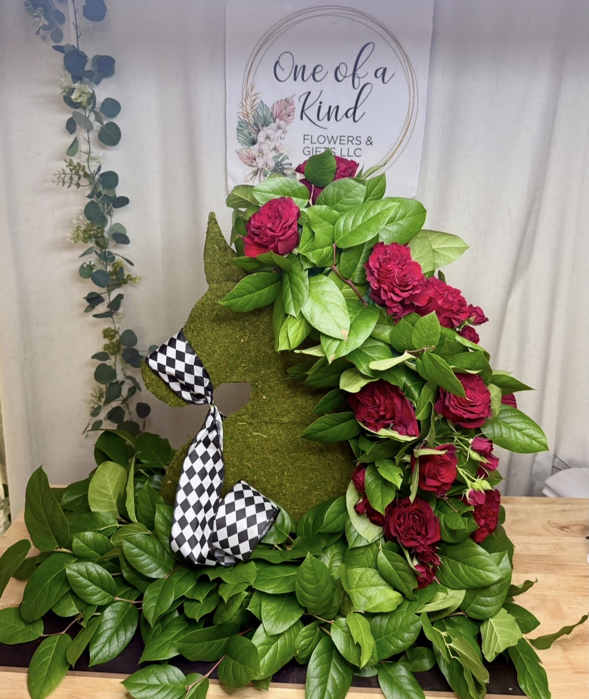 Eternal Equestrian Tribute - A unique and elegant horse-shaped floral design, created to honor a loved one with meaning, dignity, and respect. This arrangement features a natural moss-covered base, beautifully complemented by a selection of fresh red roses and premium greenery.  The design stands out for its balance of texture and volume, with cascading roses that create a striking and sophisticated visual effect. A decorative ribbon adds a distinctive finishing touch, enhancing its overall presentation.  Perfect for funeral services or memorials, this tribute symbolizes strength, freedom, and everlasting love. Each piece is handcrafted and can be customized to reflect your preferences.