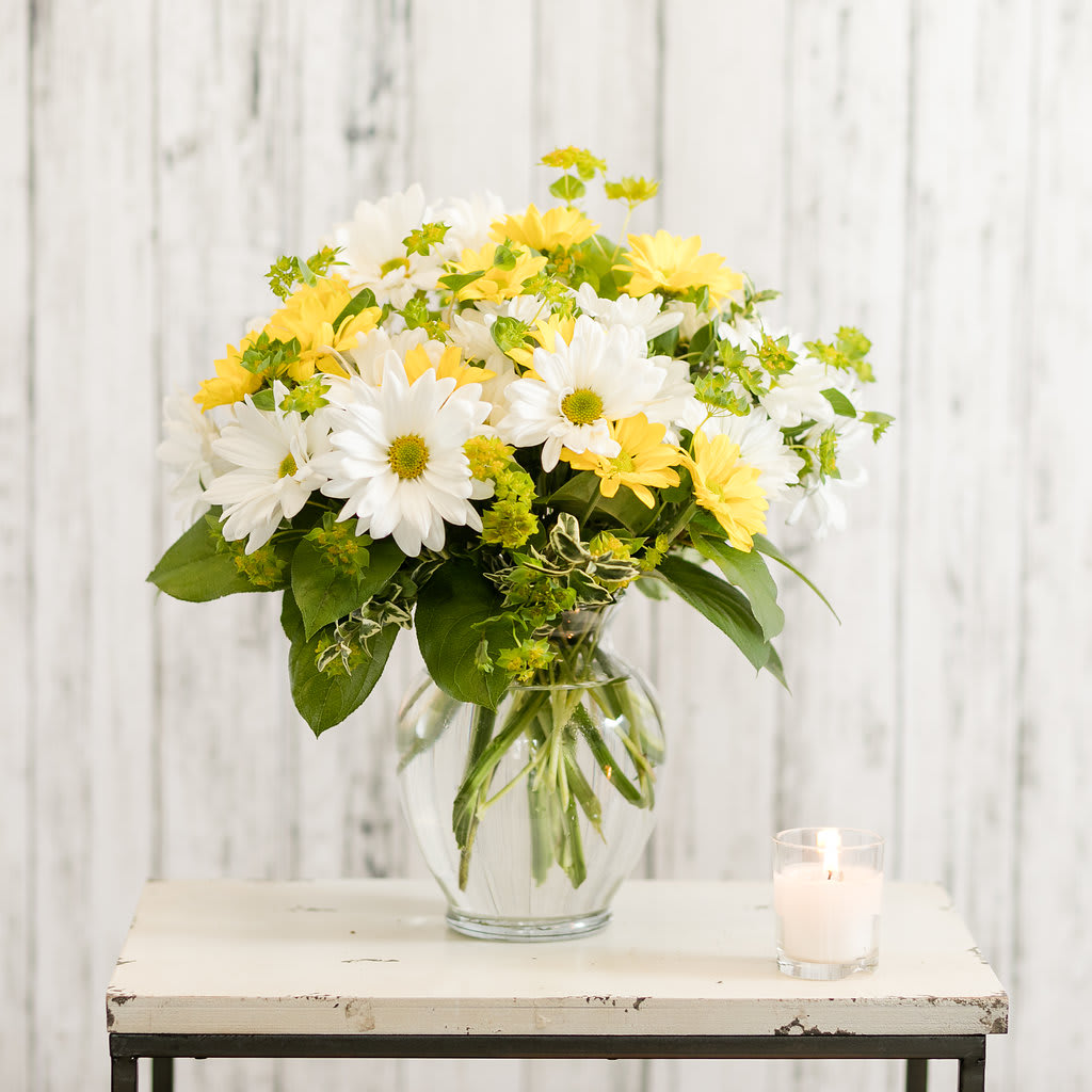 White and yellow daisy bouquet in a glass vase beside a candle