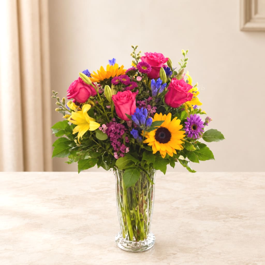 Mixed bouquet of roses, sunflowers, lilies, and purple daisies in a clear glass vase