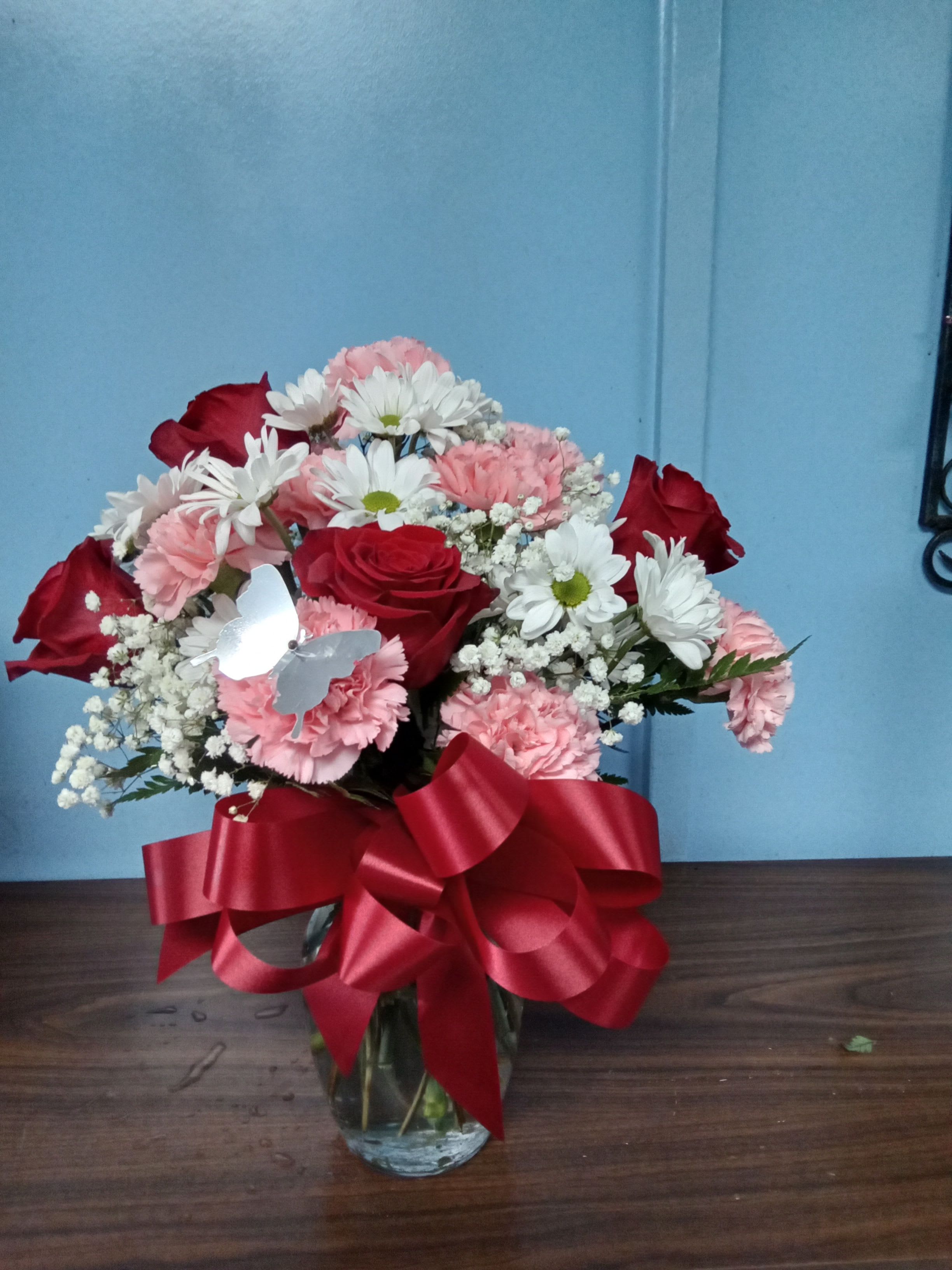Bouquet of red roses, pink carnations, and white daisies in a glass vase with a red ribbon