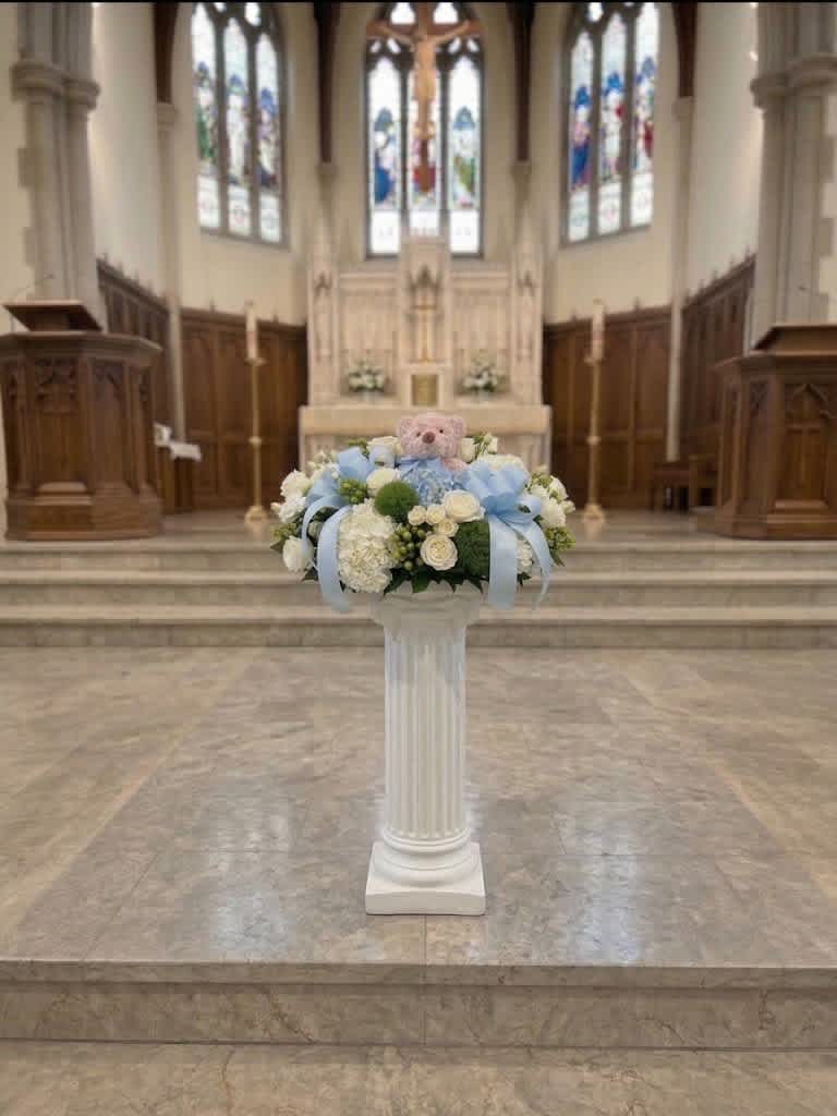 Pastel floral arrangement with a teddy bear on a white pedestal in a church