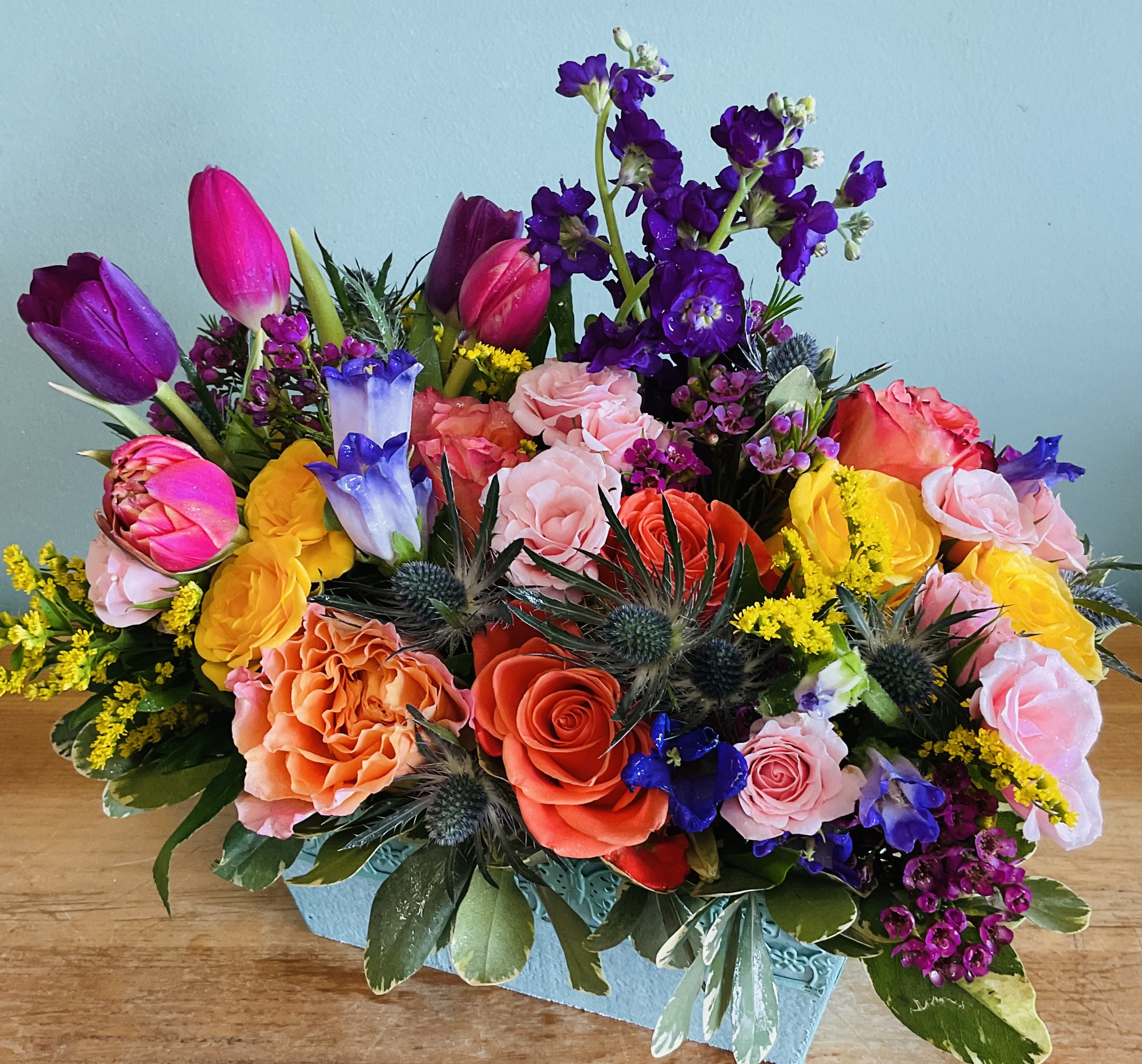 Bright Seasons - Bright Seasons opens with a burst of saturated reds, oranges, and sunny yellows-roses and upright tulips mingle with tall purple delphinium spikes for a vivid anniversary or birthday bouquet that reads like a celebration in bloom. Packed densely in a low rustic wooden box, this mixed-color flower arrangement has a bold, festive presence that makes the person you're thinking of feel truly seen-perfect for Father's Day, graduations, thank-you gifts, or a cheerful "just because" surprise.  This hand-arranged composition blends roses, tulips, dianthus and other seasonal blooms with spiky blue-green thistle-like accents, small filler flowers, silvery foliage, and glossy green leaves for layered texture and depth. The arrangement sits low and abundant in a decorative wooden container, ideal for tabletops or desks; stems and exact blooms may vary with seasonal availability, and we welcome color preferences or special requests in the order notes.  Bright Seasons also suits new baby celebrations, get-well wishes, heartfelt "I'm sorry" messages, and Grandparent's Day-any moment that calls for warmth and energy. For best results, refresh the water and trim stems as needed to keep the arrangement lively through spring, summer, and beyond.