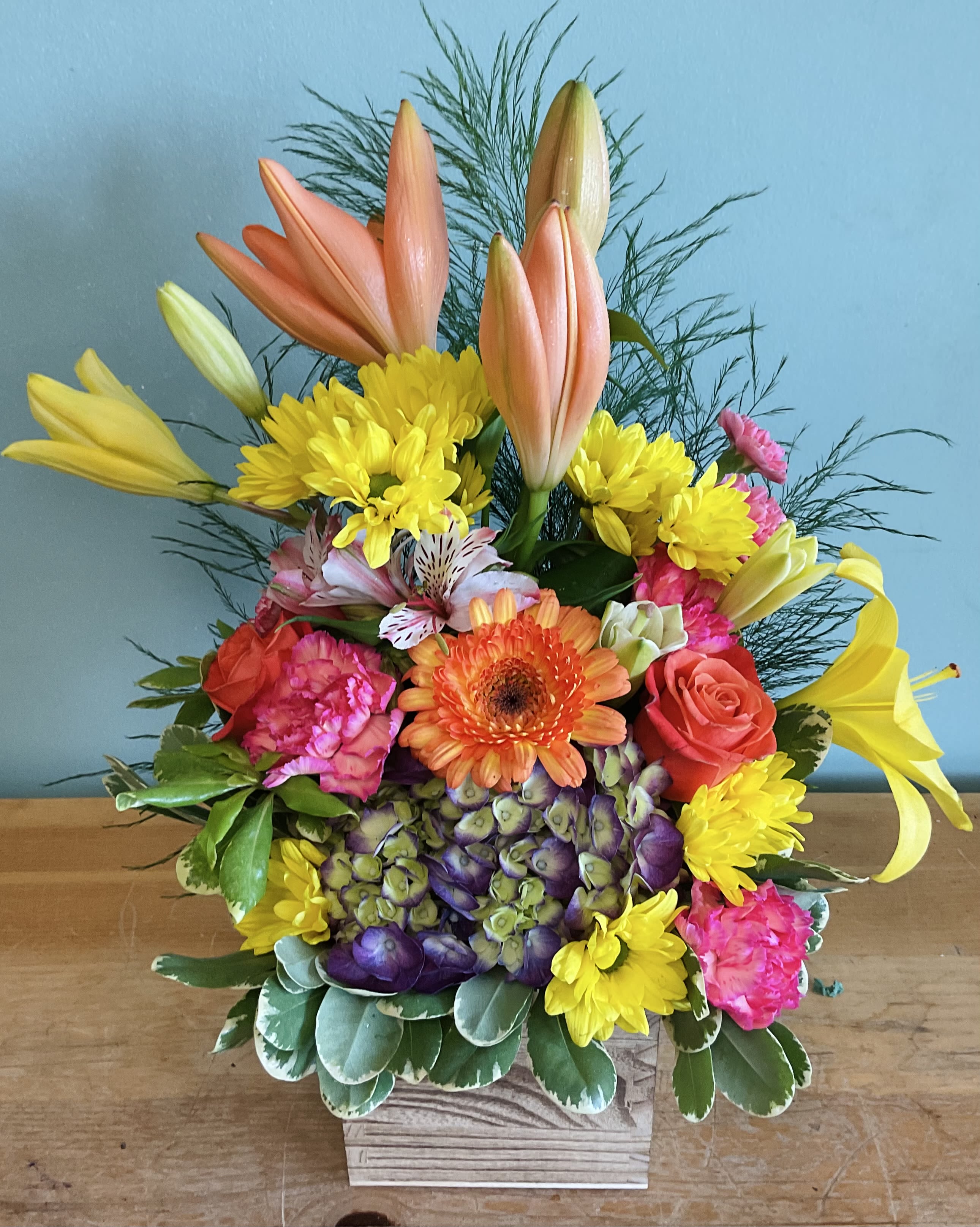 Colorful Summer Vibe - Peach and yellow lilies soar above a lush mix of pink roses, orange gerbera, vivid purple hydrangea and sunny yellow chrysanthemums in this Colorful Summer Vibe flower arrangement-perfect as a birthday bouquet, cheerful anniversary gift, or just-because centerpiece that brightens any room. The combination of warm peaches and oranges with cool purples and pinks gives the arrangement a lively, slightly tropical feel that will make the person you're thinking of truly smile.  Arranged densely in a low wooden box, the design layers tall lilies with clustered hydrangea, alstroemeria, chrysanthemums and carnations for rich texture and a full, abundant look. Feathery filler and broad variegated foliage frame the blooms and keep the composition compact enough for a dining table or office desk while still reading bold from across the room.  Send this when you want someone to feel celebrated and noticed-its saturated colors announce joy without being fussy. The wooden box provides sturdy support and easy placement; refresh with regular water changes and keep away from direct heat to extend vase life.