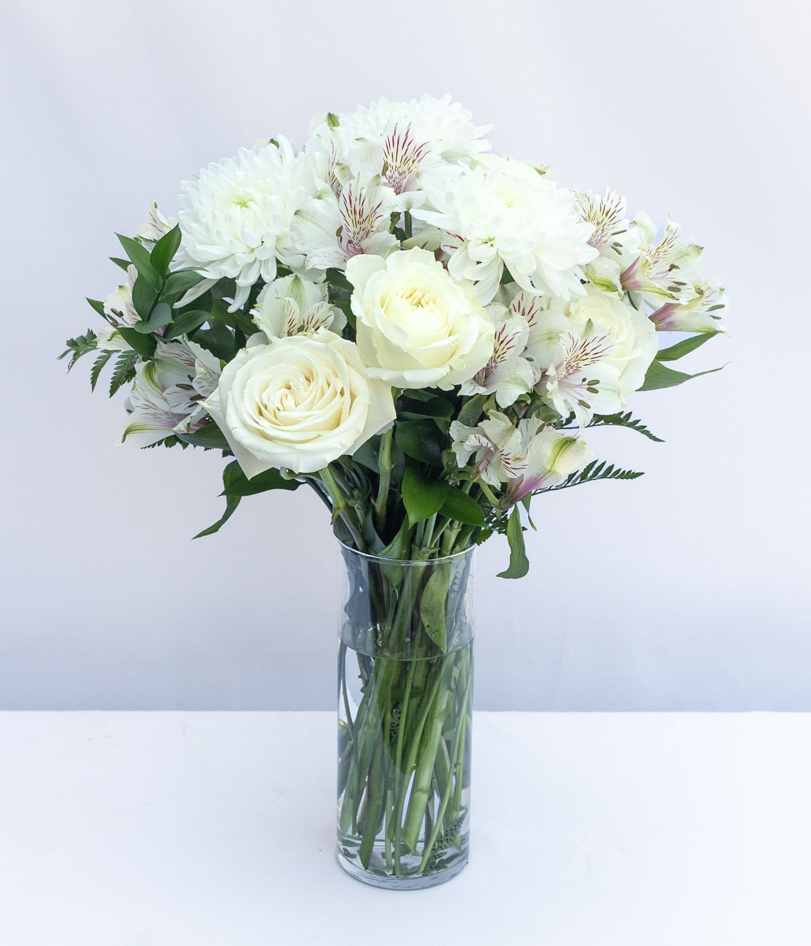 Cherished Friend Bouquet - The Cherished Friend Bouquet is designed with bright white roses and Peruvian lilies that are accented by lush greens and gorgeously arranged in a clear glass gathering vase to create a bouquet that will show how much you care.