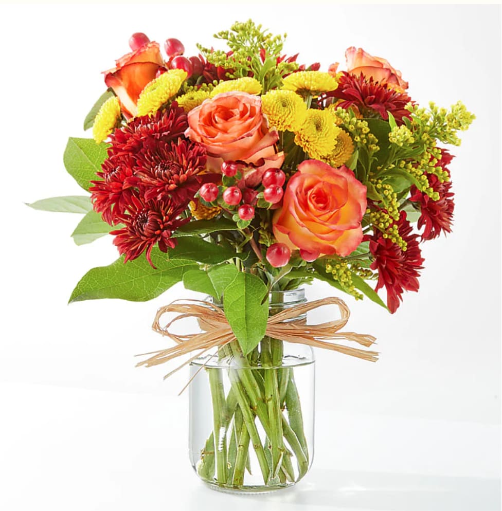 Compact bouquet of orange roses, red mums, and yellow blooms in a clear glass jar with raffia bow.