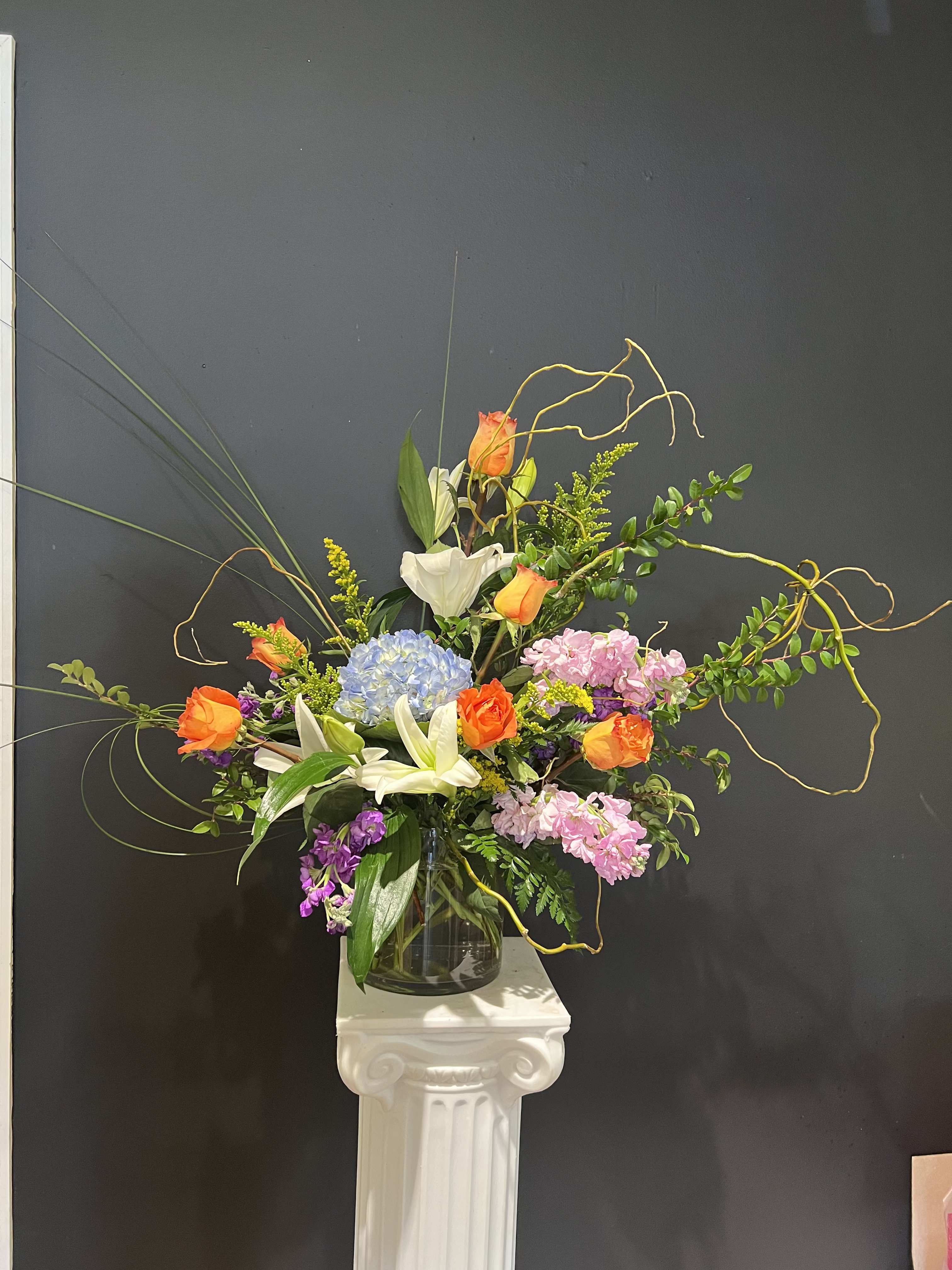 Tall mixed flower arrangement in a glass vase with orange roses, lilies, and hydrangea