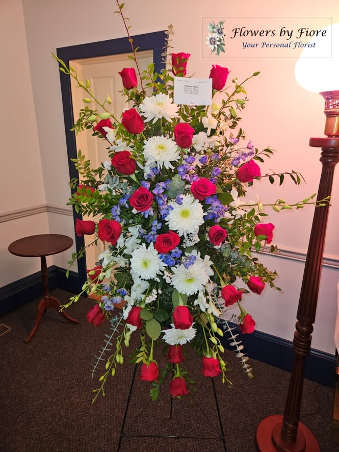 Tall standing spray of red roses, white mums, and blue flowers on a black easel
