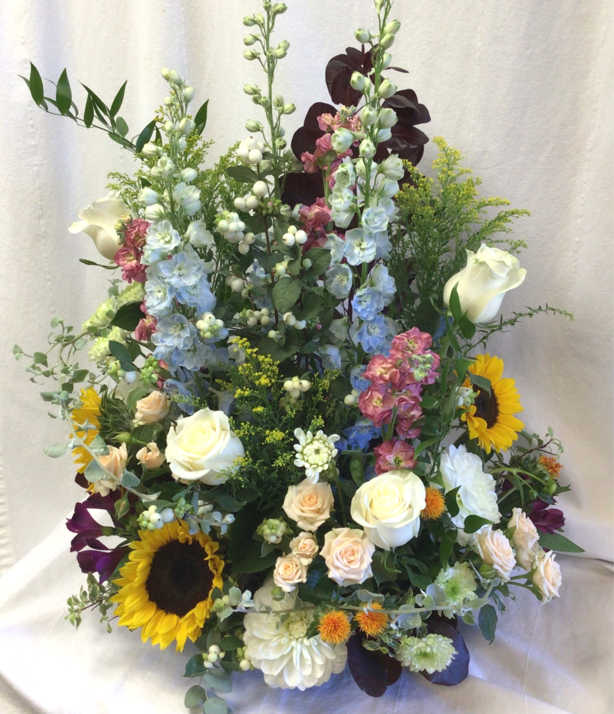  I come to the Garden - A tall, garden-style bouquet that reads like a sunlit stroll through a mixed border, perfect as a cheerful bouquet. This flower arrangement features airy height and a lush, dense base so the person you're celebrating will feel surrounded by color and texture-the kind of display that says "congratulations" or "I'm thinking of you" with warm confidence.  White roses and berries give  the arrangement an elegant, creamy center while bright sunflowers bring sunny contrast. Pale blue and pink spiked blooms (snapdragons) add vertical movement, and chrysanthemums plus small filler flowers weave the palette together in soft and vivid tones. The mix of white, pink, yellow and mixed-color blooms creates an abundant, layered look with a lively, garden-fresh scent and feel. This large arrangement arrives ready to display and make a room feel celebratory.
