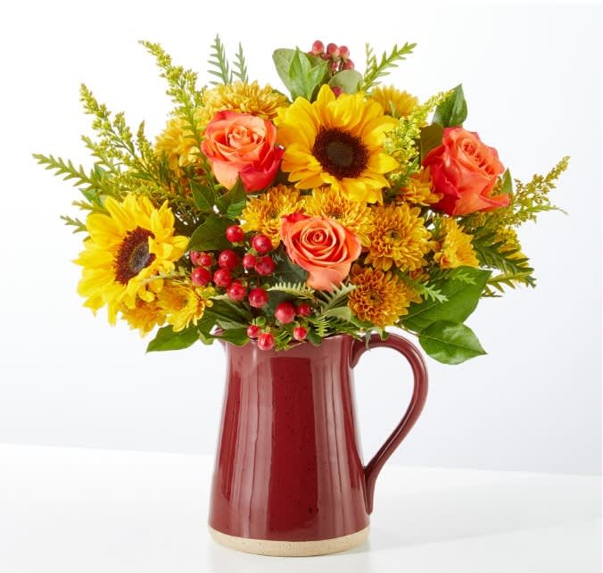 Bouquet of sunflowers, orange roses, and yellow mums in a red pitcher vase