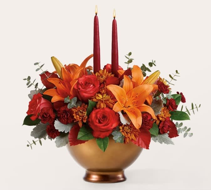 Red and orange floral arrangement in a gold bowl with two red taper candles