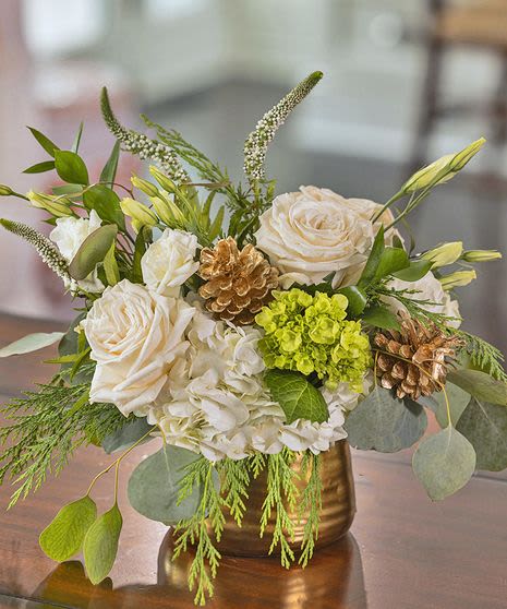 White roses and hydrangeas in a gold vase with green accents and pinecones