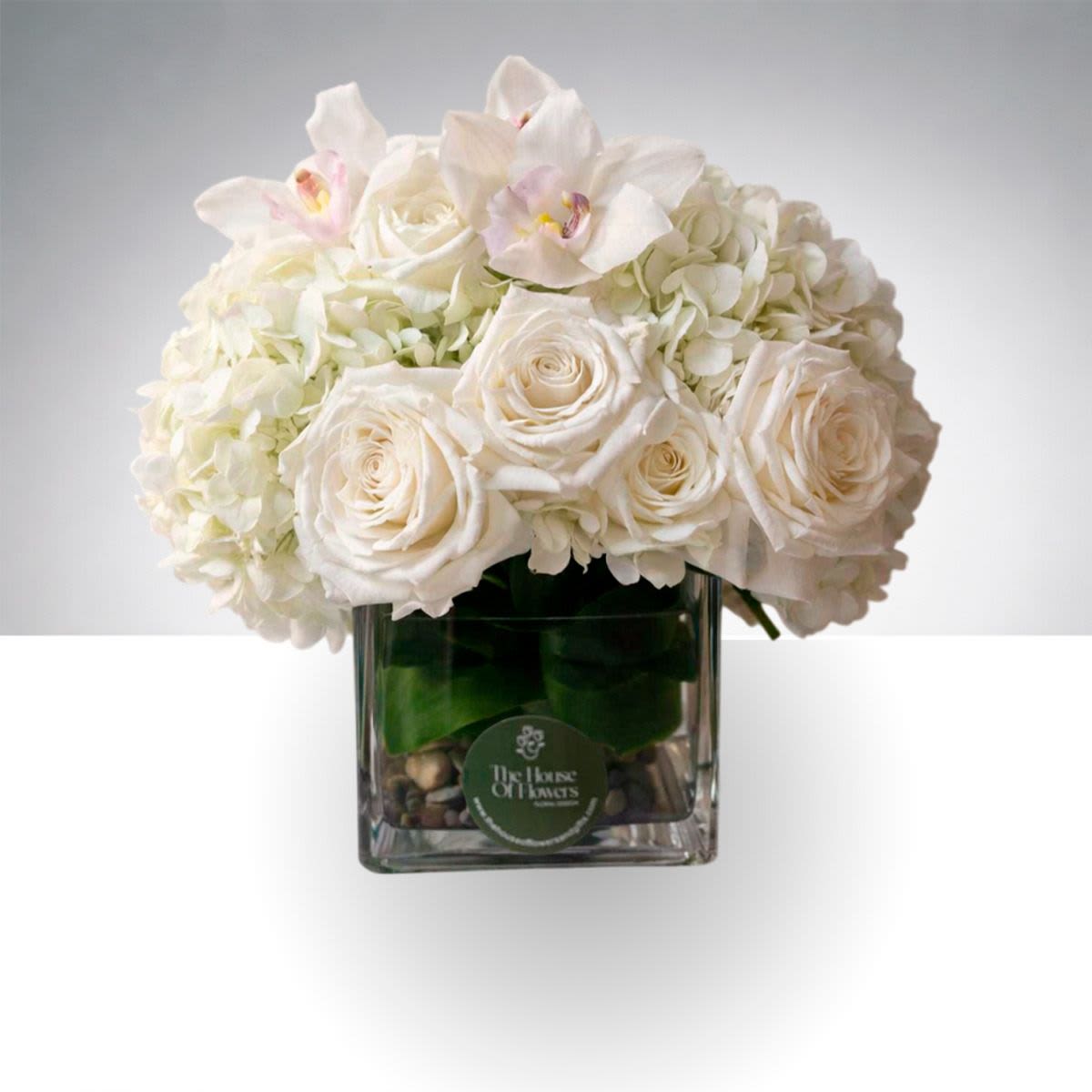 Ivory Elegance by The House of Flowers - A refined and elegant floral arrangement featuring creamy white roses, lush hydrangeas, and delicate orchid blooms, beautifully arranged in a modern glass cube with decorative stones. Fresh greenery adds natural texture and balance, creating a soft and sophisticated design.  This arrangement is perfect for home décor, celebrations, sympathy gestures, or sending a thoughtful gift that expresses grace and beauty.  A timeless white floral arrangement designed to bring elegance and serenity to any occasion.