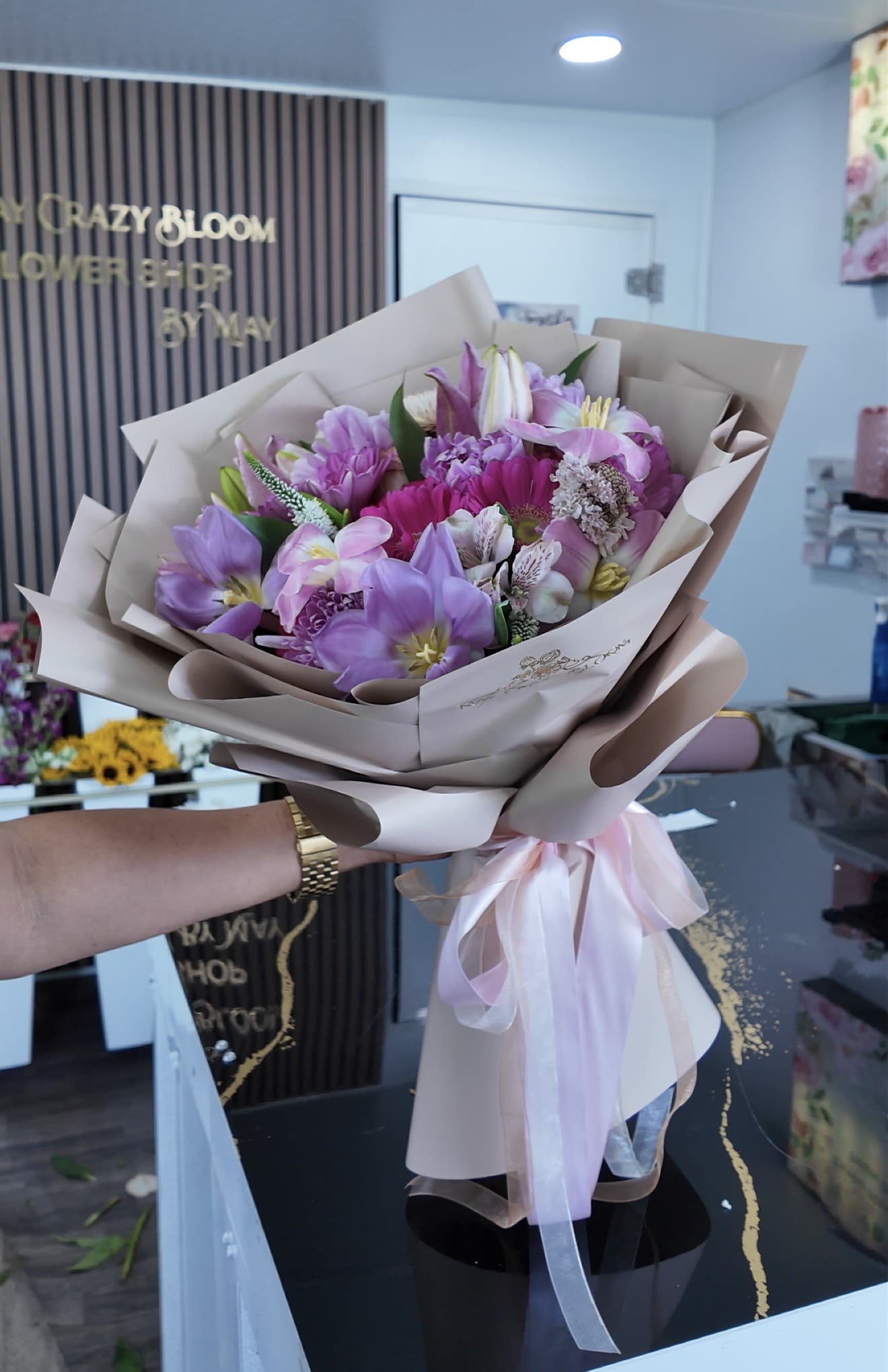 Pinks - Clouds is a soft, romantic handheld flower arrangement perfect as a birthday bouquet, anniversary surprise, or a thoughtful thank-you gift. The mix of pink, lavender, white and peach tones-anchored by lilies, tulips, gerbera daisies, carnations, alstroemeria and clustered chrysanthemums-creates a gentle, cloud-like impression that will make the person you're thinking of feel seen and celebrated.  This florist-tied bouquet is arranged in a full, rounded shape and wrapped in layered beige paper with satin and pink ribbon finishes for a polished presentation. Expect a lively yet delicate texture from the larger daisy faces and lily blooms balanced by smaller filler flowers; the color palette reads fresh and springlike while remaining versatile year-round.  Choose this arrangement when you want to send a cheerful, feminine statement that reads both everyday and special-ideal for moms, friends, or partners. The hand-tied format arrives ready to place in a vase; for best longevity, trim stems and refresh water every 2-3 days.
