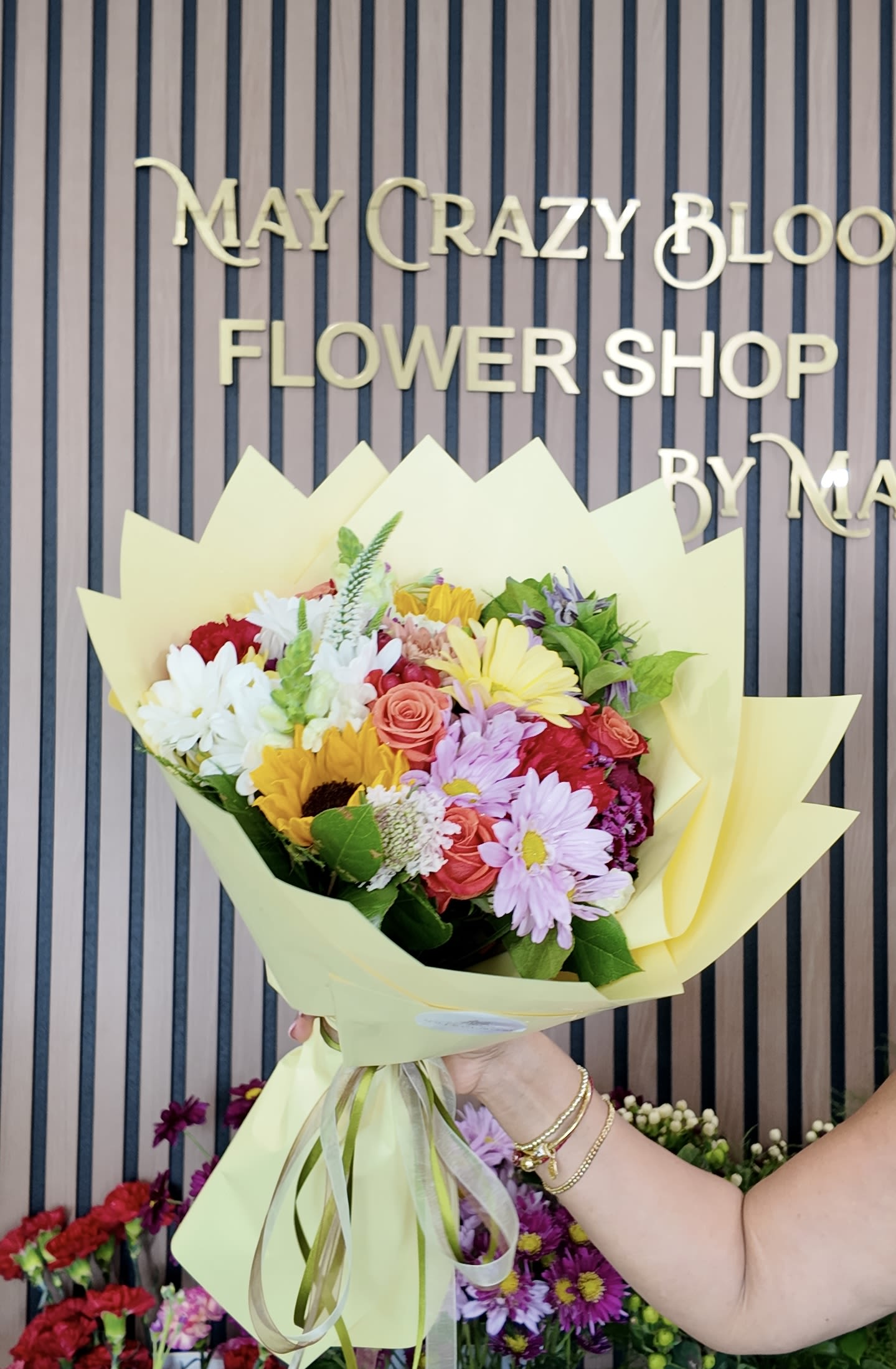 Rain - A lively mixed-color bouquet that reads like sunshine in your hands, Rain is a cheerful flower arrangement perfect for birthdays, congratulations, or "just because" moments. The hand-tied shape and soft pale yellow wrapping give this bouquet a bright, gift-ready look that makes the person you're thinking of feel celebrated and truly seen.  This arrangement mixes roses, daisies, sunflowers, chrysanthemums, snapdragons, carnations and other seasonal blooms in a full, rounded cluster. Expect pink and coral roses, white and pink daisy-like blooms, yellow sunflowers and accents of lavender and red chrysanthemums, all wrapped in pale yellow paper with a matching ribbon for a soft, joyful presentation. The contrast between vivid petals and the gentle wrap creates a lively, slightly romantic mood.  Rain is ideal for sending warm congratulations, birthday wishes, or an everyday pick-me-up that reads as thoughtful and vibrant. Care is simple: place stems in fresh water upon arrival, recut stems at an angle every few days, and keep the bouquet out of direct heat to extend its life.