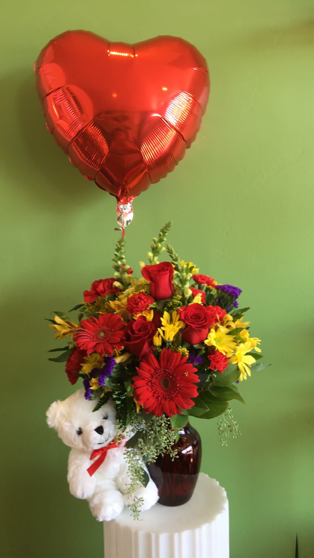Red heart balloon above a mixed flower arrangement with a teddy bear