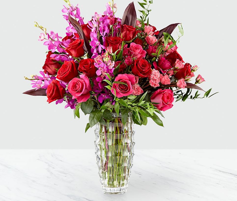Heart's Wishes™ Luxury Bouquet by Interflora™- VASE INCLUDED - Deeply devoted to claiming your special recipient's heart with a bold mix of color saturated in it's finest form, this bouquet speaks to everlasting romance with each and every stem. Rich red roses mingle with the eye-catching hot pinks of Mokara Orchids, roses, and spray roses that help to convey your heart's every wish. Finished with lush green accents to delight the eye and make each bloom pop in it's intended way, this luxury bouquet arrives in a clear faceted glass vase lined with exotic red ti leaves to add even further interest to this already opulent design style. A gift of love that will surely touch their heart. IMPRESSIVE bouquet is Approx. 24"H x 26"W. STUNNING bouquet is Approx. 25"H x 27"W.