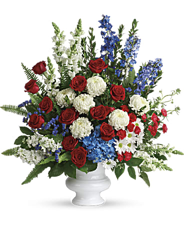 Large red, white, and blue flower arrangement with roses, mums, and delphinium in a white pedestal vase