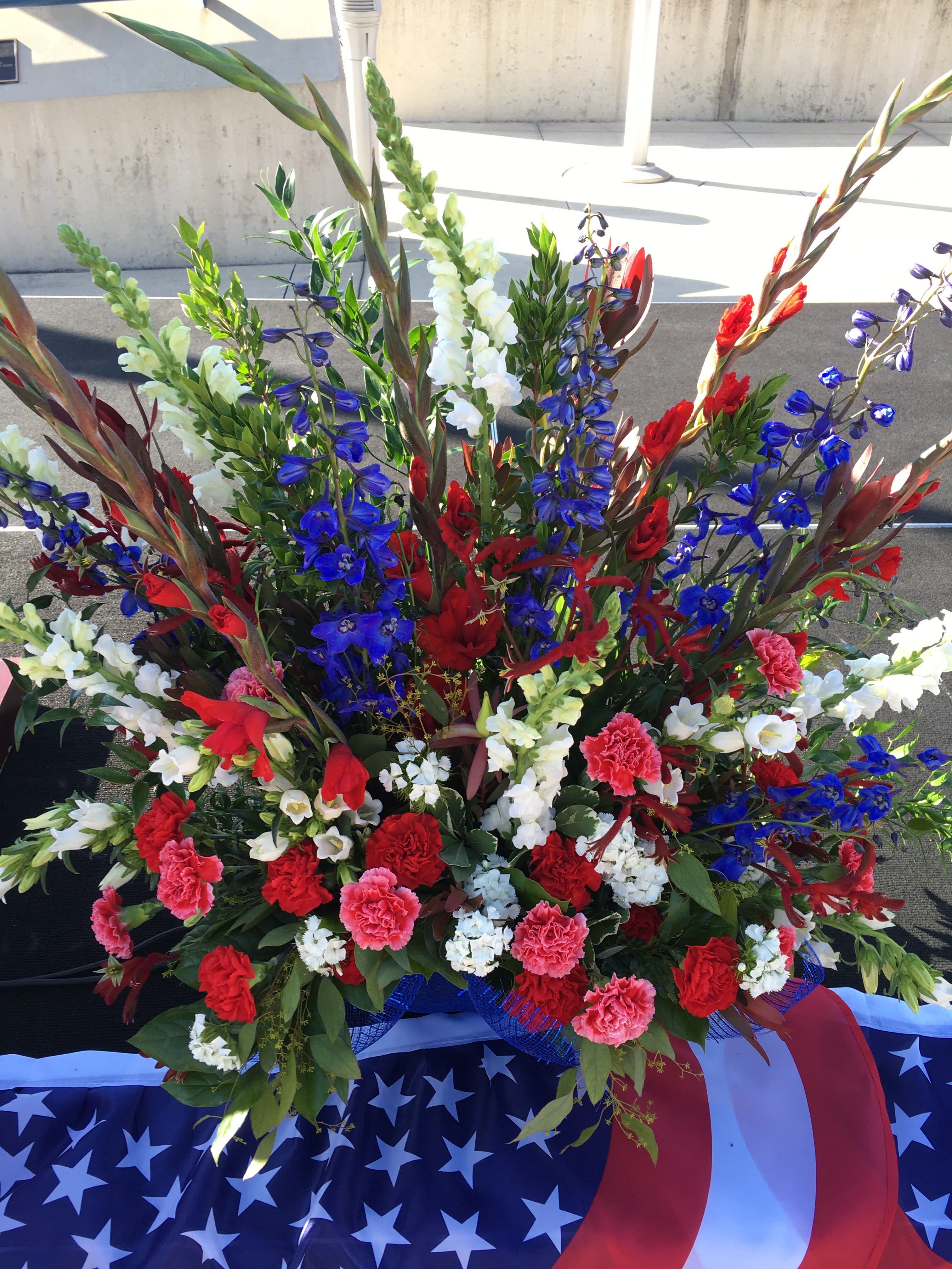 Large Patriotic mache basket - Brilliant red, white, and blue flowers in a fan shaped spray design. This is a floral foam design in a papier-mâché basket meant for a stage, lawn, church or hall ceremony.  Selection will vary.  Approximately 3 feet tall and 4 feet wide. Celebrate your veteran with this beautiful upbeat creation. 